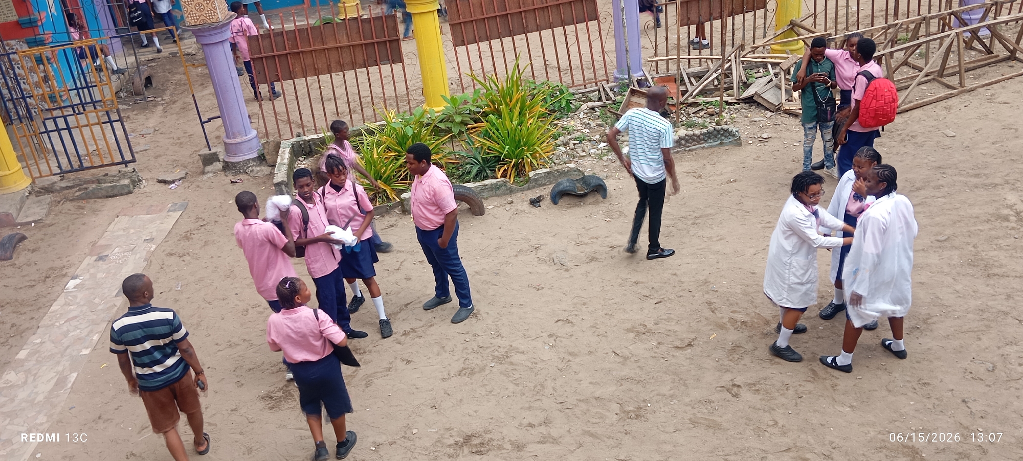 This image depicts an outdoor scene in a sandy, urban environment that appears to be a schoolyard or recreational area. Several individuals, likely students based on their attire, are present. The majority are wearing pink uniforms with dark blue skirts or trousers, and some have white lab coats over their uniforms. One adult male in a striped t-shirt and brown shorts is visible, as is another male in a green t-shirt and ripped jeans.
The background features a light brown fence, colorful metal playground equipment, and scattered debris including wooden structures and discarded tires. The ground is primarily composed of sand, with some patches of concrete or paving. The lighting suggests it is daytime. The timestamp at the bottom right indicates "06/15/2026 13:07", which could place the image in Lagos, Nigeria, given the context of the prompt.
Specific actions include students conversing, one student on their phone, and another carrying white material. The overall atmosphere suggests a casual gathering or transition period within the school day.