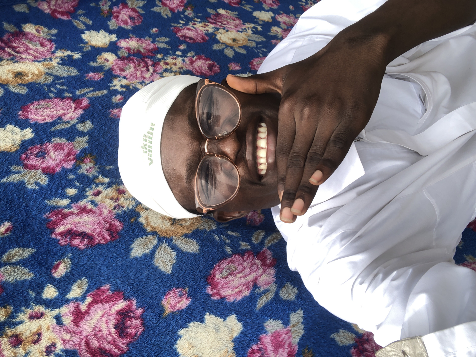 A medium shot captures a young Black man with dark skin, wearing a white cap and white shirt, from the chest up. He has on large, round, rose-gold rimmed glasses. The man is smiling broadly, revealing his teeth, and has his right hand resting on his chin. His thumb is extended upward and his fingers are curved, resting on his cheek. The background is a blue tapestry with a floral pattern of pink and yellow flowers and green leaves. The man's white cap has light green text on it that reads "Ike Vanna." The lighting suggests an outdoor setting, possibly under direct sunlight, given the strong contrast and shadows on his face and clothing. The scene appears to be in Ado, Nigeria, based on the provided location context.