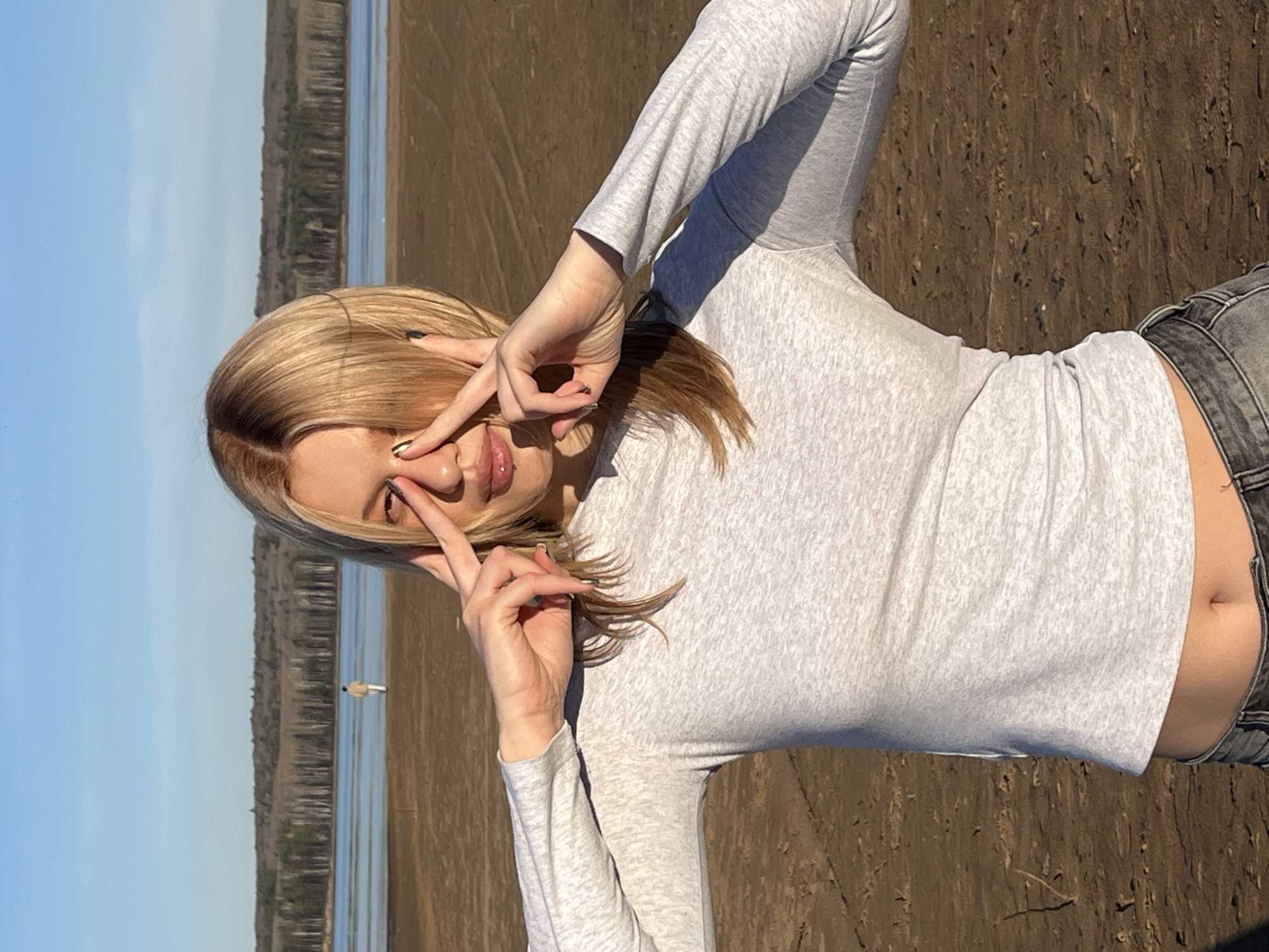 A young woman stands outside in Chaikovsky, Russia. She has light blonde hair with dark roots and is wearing a light gray long-sleeve shirt and gray jeans. She is making a peace sign with both hands in front of her face. In the background, a lake or large river is visible, with a shoreline of trees and some buildings in the distance. The ground is brown and appears to be dirt or sand. The sky is light blue.