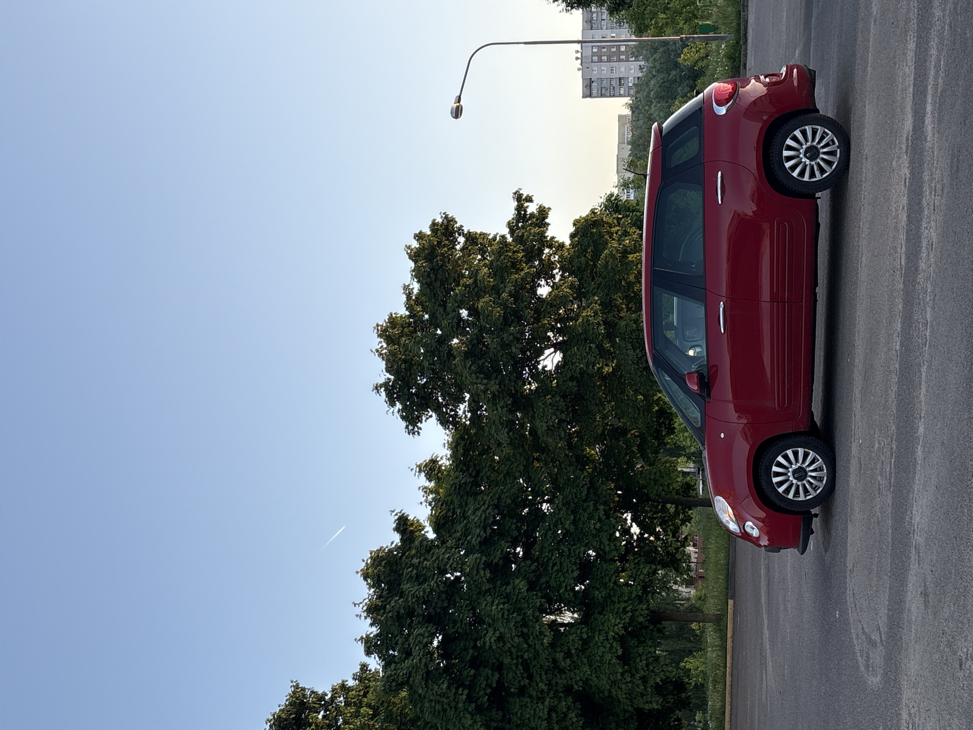 The image captures a red Fiat 500L parked on an asphalt surface, likely a parking lot or street, in Minsk, Belarus. A large, lush green tree stands beside the car, contrasting with the clear, blue sky. A tall streetlight is visible in the background, as is an apartment building. The lighting suggests it is either early morning or late afternoon. The car is the main subject, with its vibrant red color and distinctive shape. The presence of the tree and building help to further place the scene in Minsk. There are no people visible in the frame.