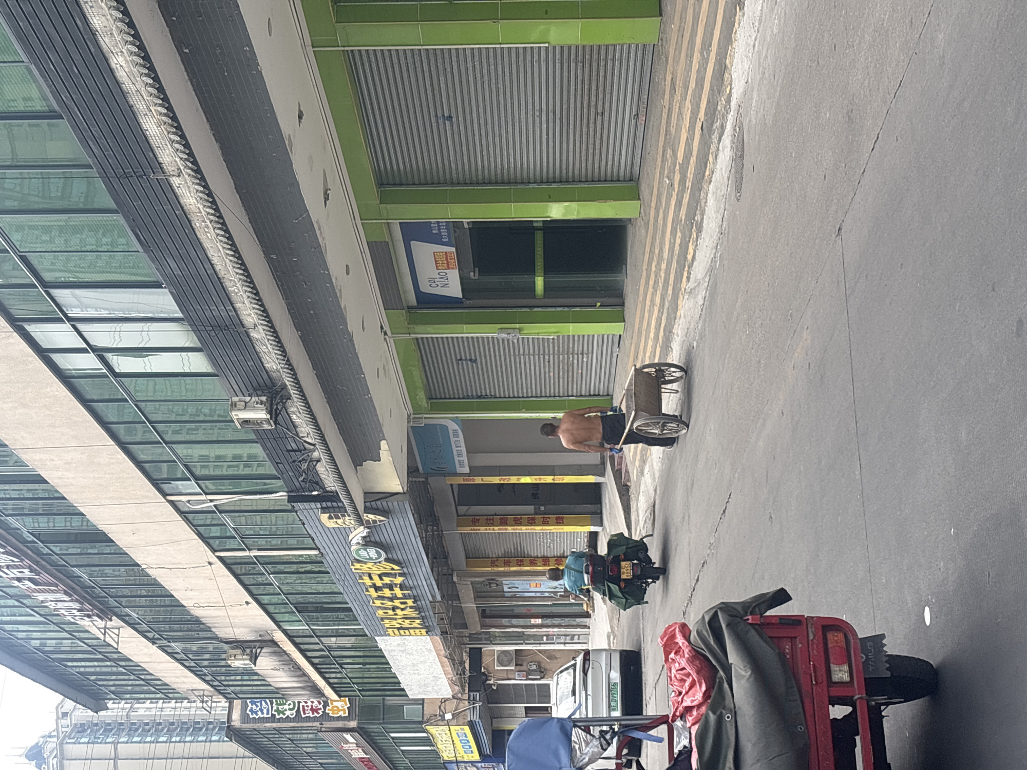 The image shows a street scene in Nanhai District, China, with a focus on a row of shops. In the foreground, a red three-wheeled vehicle is parked, partially covering a large green tarp and a red cloth. A person is riding a motorcycle with saddlebags, heading away from the camera. Another shirtless man is walking towards the camera, pushing a handcart with a wheelbarrow on it. The shops have green tiled facades and metal roller shutters, indicating they are likely closed. Some shops have signage with Chinese characters and logos. The street is paved and appears to be an urban area. The overall impression is of a commercial street on an overcast day.