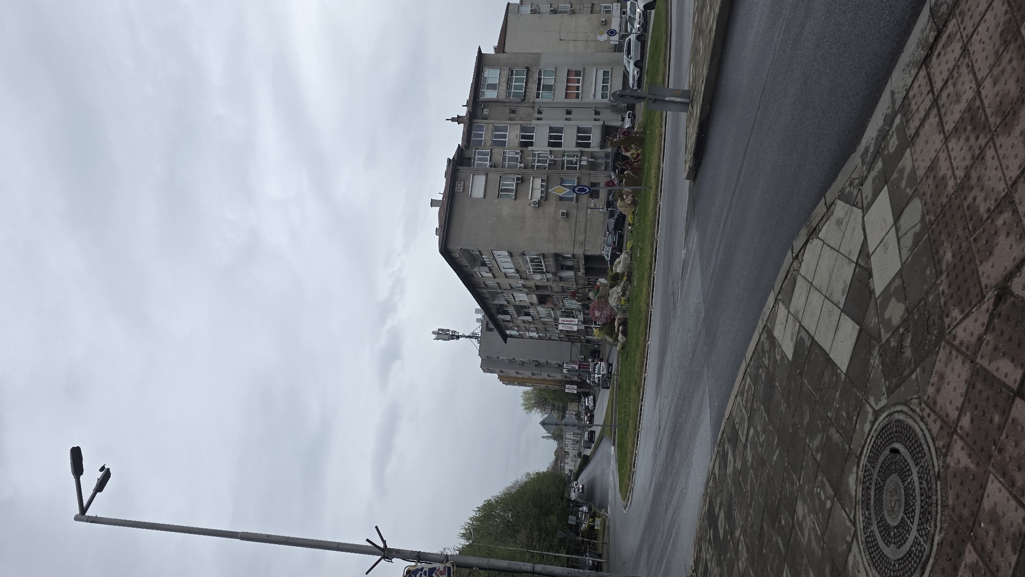 The image depicts a street scene in Blagoevgrad, Bulgaria, under overcast skies. Several multi-story residential buildings line the street, constructed with concrete facades and featuring numerous windows. A cellular antenna is mounted on one of the buildings. Parked cars are visible along the curb. The street itself is paved with asphalt, appearing wet, and is bordered by a green strip of grass. A sidewalk made of patterned tiles runs along the edge of the street. A lamppost stands in the foreground. Traffic signs are present, though partially obscured.