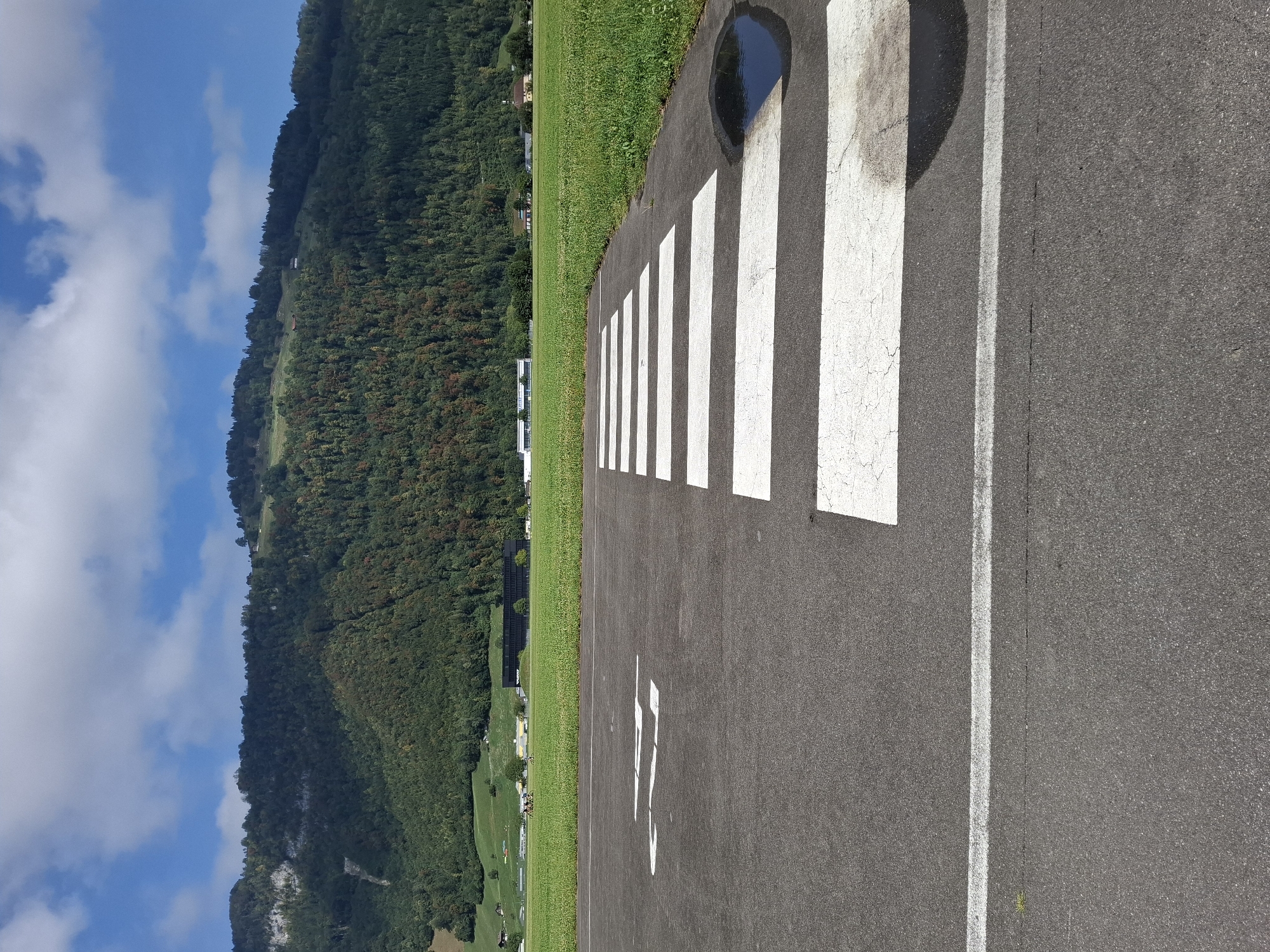 The image shows a runway at the Buochs Airport in Buochs, Switzerland. In the foreground, there is a tarmac surface with white markings, including a runway designation and a series of white stripes that appear to be part of a pedestrian crossing or a painted line. A small puddle of water is visible on the tarmac near the white stripes. Beyond the runway, a vast green field stretches towards a densely wooded mountain range under a bright blue sky with scattered white clouds. Several buildings and structures are visible in the distance, nestled at the base of the mountains and scattered across the green landscape. The scene suggests a clear, sunny day in an outdoor, possibly rural, environment.