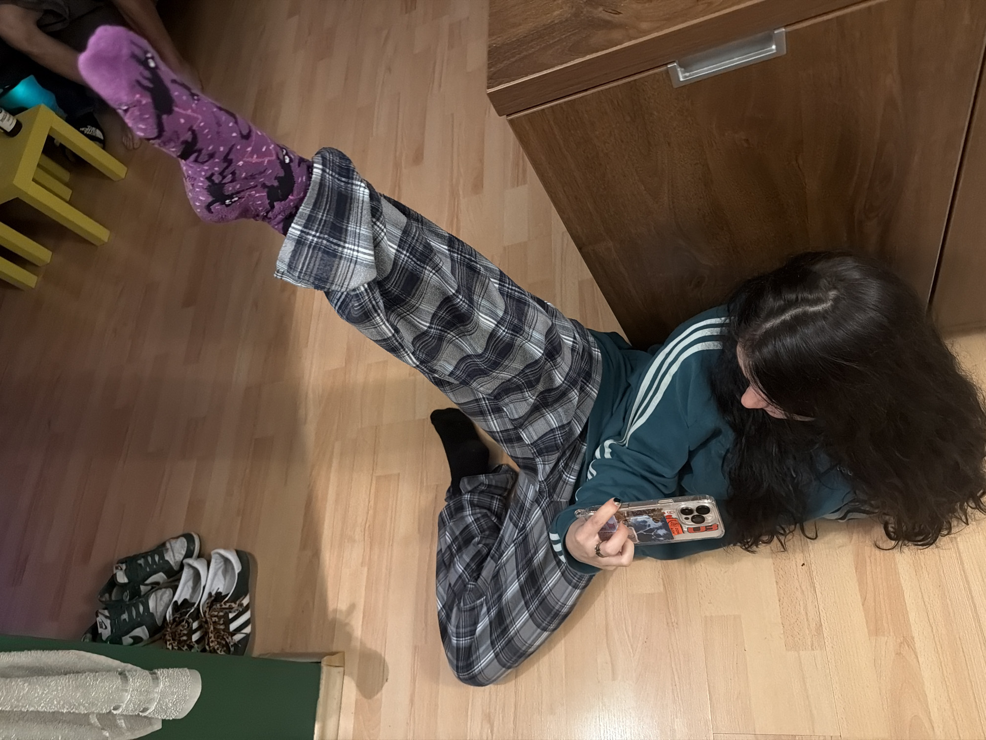 This indoor photograph, taken in Sofia, Bulgaria, captures a casual scene featuring two individuals relaxing. The primary subject is a person with long, dark, wavy hair, seen from an overhead perspective, lying on their left side on a light wooden floor. They are wearing a dark teal tracksuit top with white stripes on the sleeves and comfortable plaid trousers in shades of dark gray, black, and white. Their focus is entirely on a smartphone held in their left hand, which has a clear case displaying a design that includes the text "SPOT." A ring is visible on their left hand, and their fingernails are painted dark. Their left foot is propped up, revealing a vivid purple sock patterned with black shapes resembling bats or spiders and small white dots. Their right foot is clad in a plain black sock.
In the upper-left corner, a second person is partially visible, seated on a bright yellow, two-tiered stool. Only their lower legs and feet are seen; one foot is bare, while the other wears a dark shoe or sock. A bottle is also present on the yellow stool. The background features a tall wooden cabinet with a silver handle, and the wooden flooring extends throughout the visible area. In the bottom-left corner, three pairs of sneakers are neatly arranged on the floor, including one pair with distinct white and black stripes and leopard-print laces, likely Adidas brand, matching the visible stripes on the primary subject's top. A white towel or textured fabric is partially visible on a green surface in the same corner. The scene conveys a relaxed, domestic atmosphere, with the time of day being indeterminate due to indoor lighting.