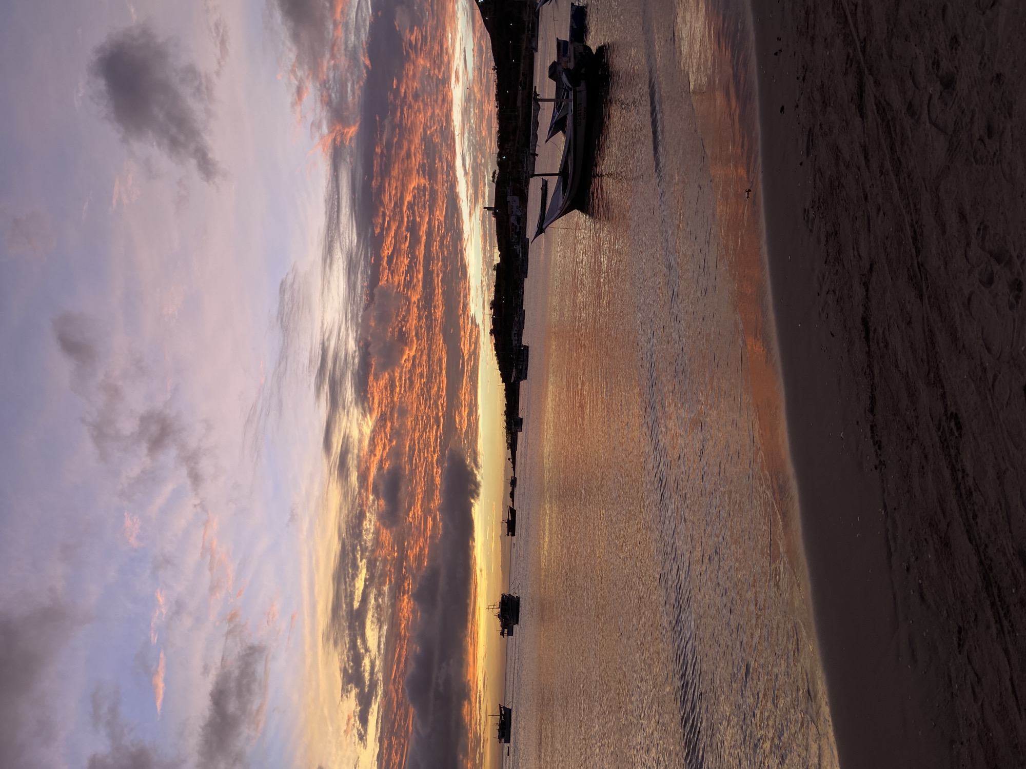 This is a photograph of a sunset over the bay of Juan Griego, Venezuela. The sky is filled with dramatic clouds painted in hues of orange, pink, and purple. The water reflects the colorful sky, creating a serene and vibrant scene. Several boats are anchored in the bay, their dark silhouettes contrasting with the illuminated sky and water. The foreground shows a sandy beach with some tire tracks. The overall atmosphere is peaceful and picturesque.