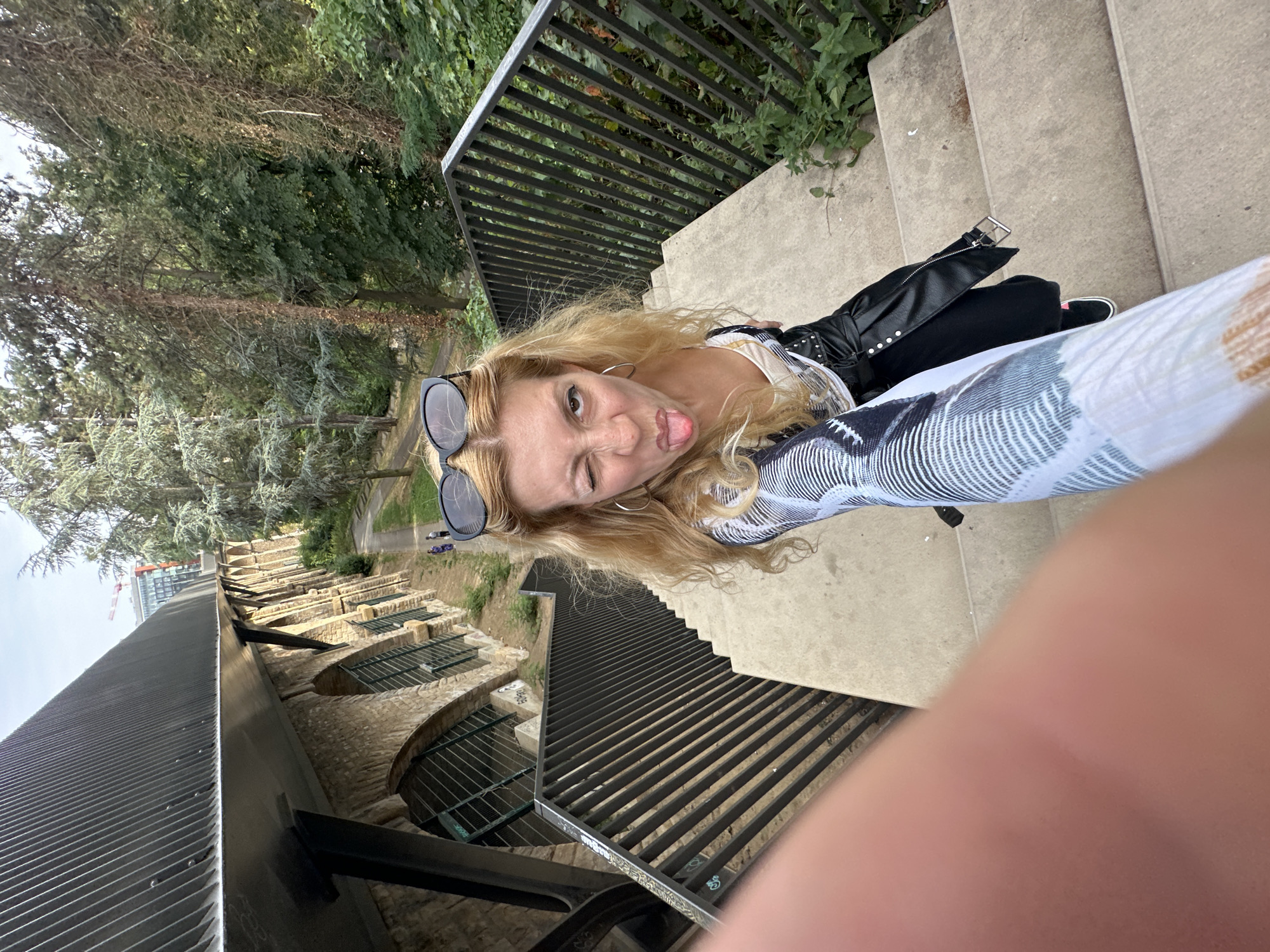 A blonde woman is taking a selfie on a concrete staircase in a park in Luxembourg City. She is wearing sunglasses on her head, a white top with a patterned jacket, and a black leather skirt. She is sticking her tongue out and winking at the camera. The staircase has a black metal railing on the right side. In the background, there is a stone building with arched windows, trees, and a distant building under construction with a crane. The lighting suggests it is daytime.