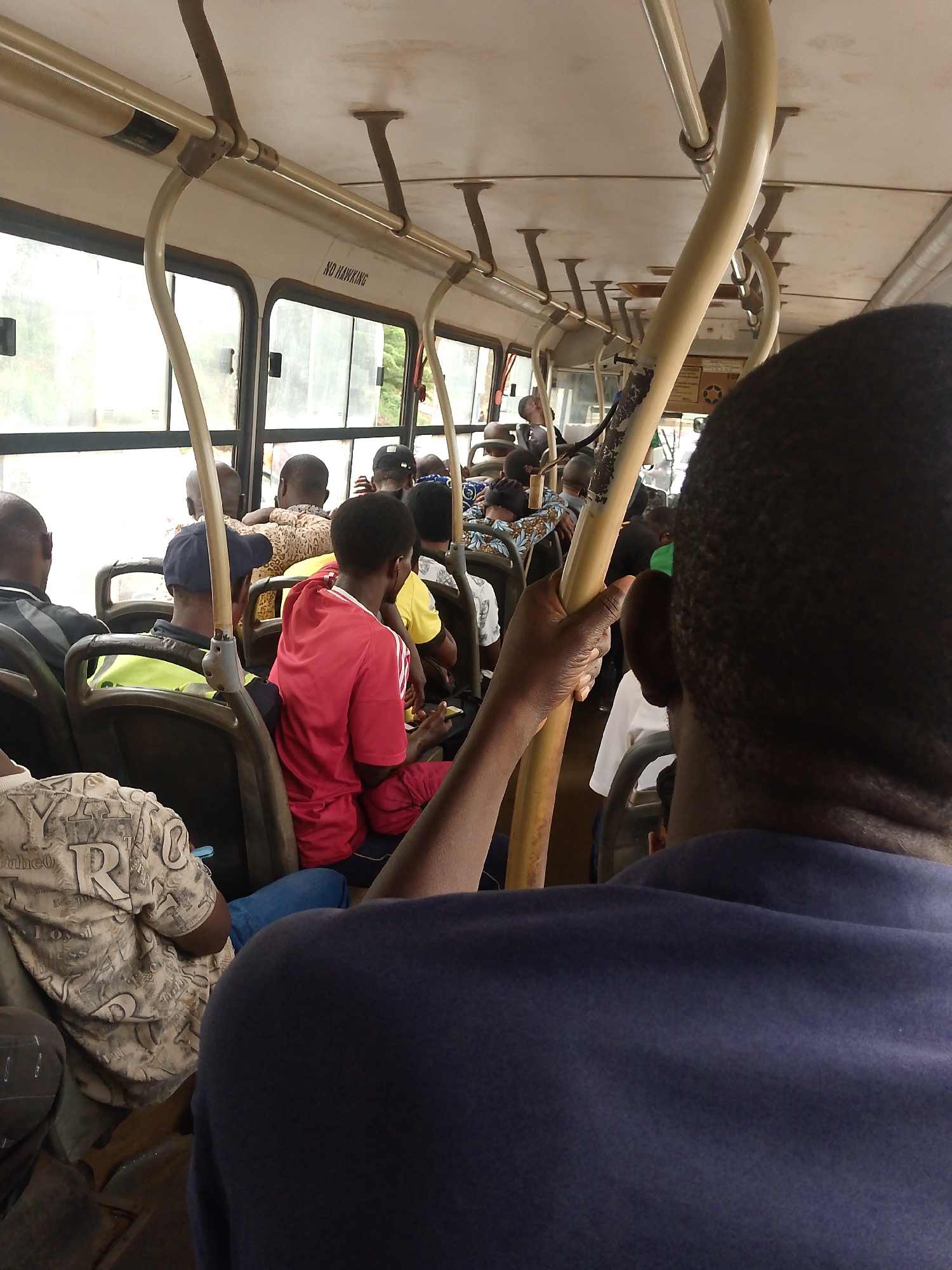The image depicts the interior of a public bus in Masaka, Nigeria, with passengers seated. The camera is positioned from the perspective of a passenger in the foreground, facing forward. The bus appears to be crowded, with people visible in the seats and along the aisles. The interior features beige-colored panels on the ceiling and windows that offer a view of greenery outside. Several yellow support poles are strategically placed throughout the bus to assist passengers. A sign reading "NO HAWKING" is visible on one of the interior panels above the windows. Passengers are diverse in age and attire, with some wearing casual clothing and others in more formal shirts. One passenger in the middle ground is wearing a red t-shirt, and another in the foreground is wearing a navy blue shirt. The overall atmosphere suggests a typical commute in a busy urban environment.