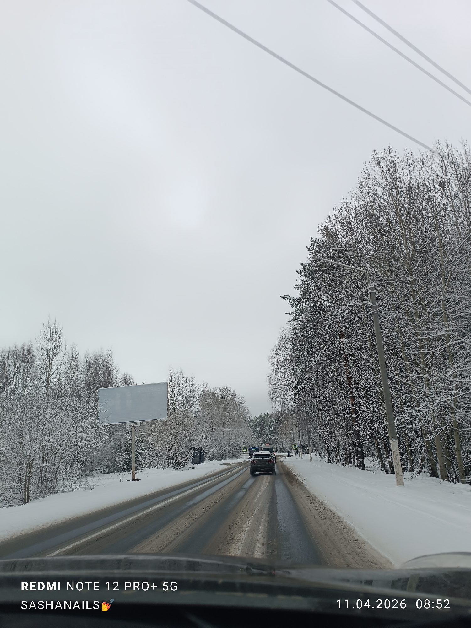 A landscape view from inside a vehicle driving on a road in the city of Chaikovsky, Russia on April 11, 2026 at 08:52. The road is wet and partially covered with snow. Snow-covered trees and bushes line both sides of the road. A blank billboard is visible on the left. Another vehicle is in front on the same lane, and overhead power lines stretch across the sky. The sky is overcast. The photograph was taken with a Redmi Note 12 Pro+ 5G.