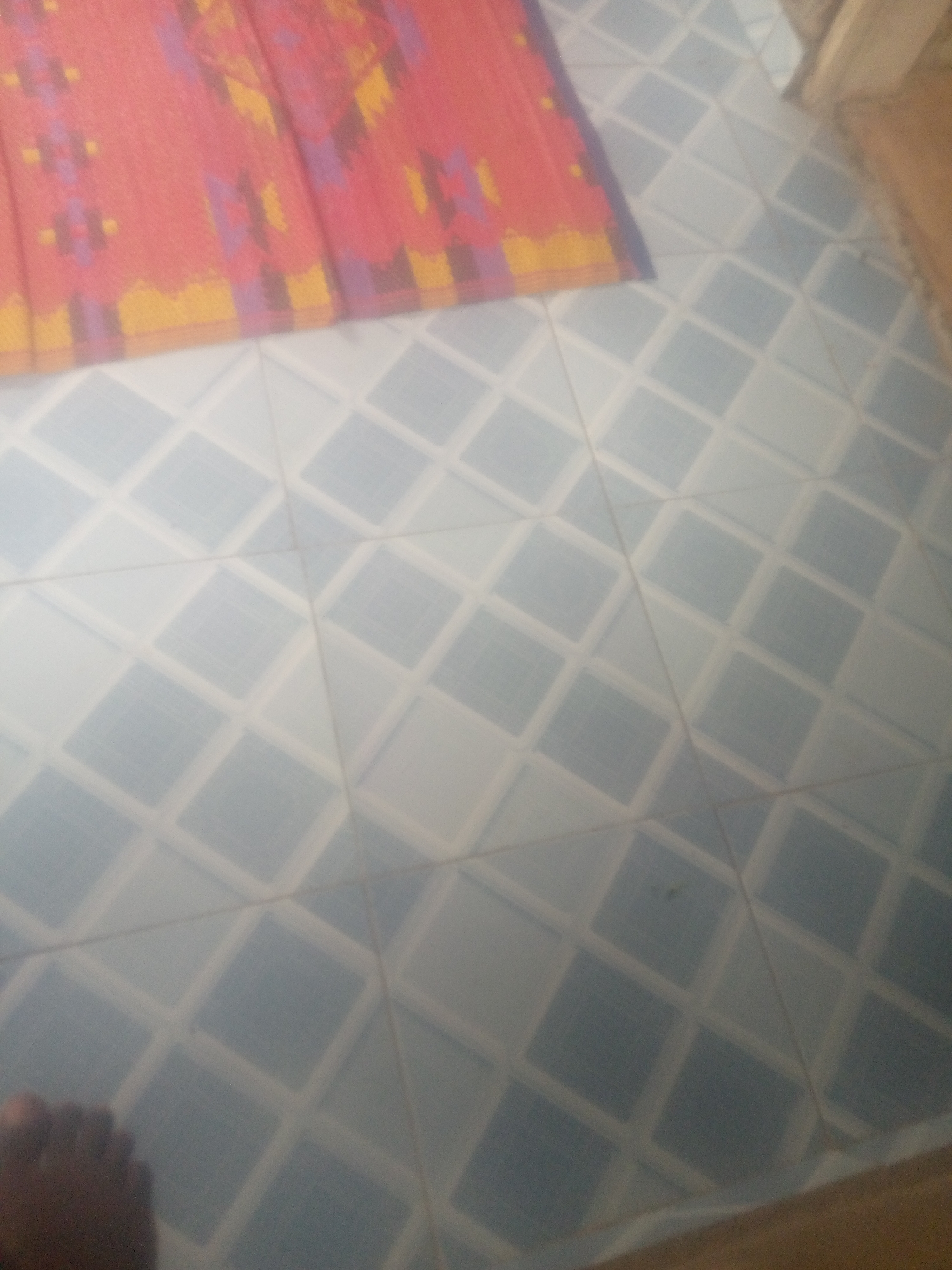 A high-angle, medium shot captures a close-up view of a floor adorned with diamond-shaped tiles. The tiles are primarily a light blue, bordered by white grout that forms a diamond pattern. In the upper left corner of the image, a portion of a red, yellow, and purple patterned rug or fabric is visible, partially obscuring the tiled floor. The lower left corner of the image reveals the partial view of a dark-skinned foot, with visible toes, resting on the tiles. The lighting suggests an indoor environment, with no specific environmental features indicating a city location.