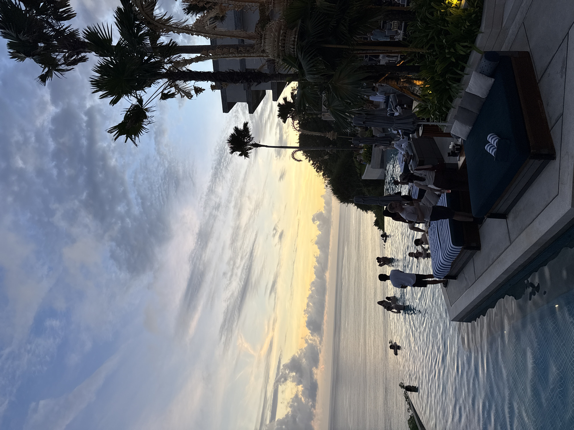 The image depicts a twilight scene at an infinity pool in Kuta Selatan, Indonesia. The pool extends towards a light-streaked sky over the ocean. Several people are in the pool, some standing near the edge. A partially visible lounge bed with striped pillows and cushions sits on the tiled pool deck. Palm trees and building structures are visible above the pool area.
