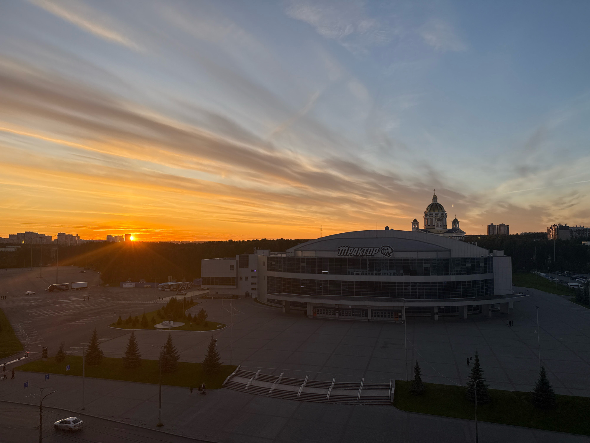 This is an aerial shot of the Traktor Ice Arena in Chelyabinsk, Russia, at sunset. The sun is setting on the left side of the frame, casting a warm orange glow across the sky and illuminating the clouds with streaks of pink and yellow. The ice arena, a modern structure with a curved glass facade, dominates the foreground and middle ground. The text "ТРАКТОР" (TRAKTOR) is visible on the upper part of the arena. In the background, a domed building, possibly a church or other religious institution, stands against the darkening sky, along with several residential buildings and trees. The surrounding area appears to be a large, open paved space, likely a parking lot or plaza, with some parked vehicles, including trucks and buses, and a few individuals walking around. The overall scene conveys a sense of an urban environment at dusk, with a blend of modern architecture and natural light.