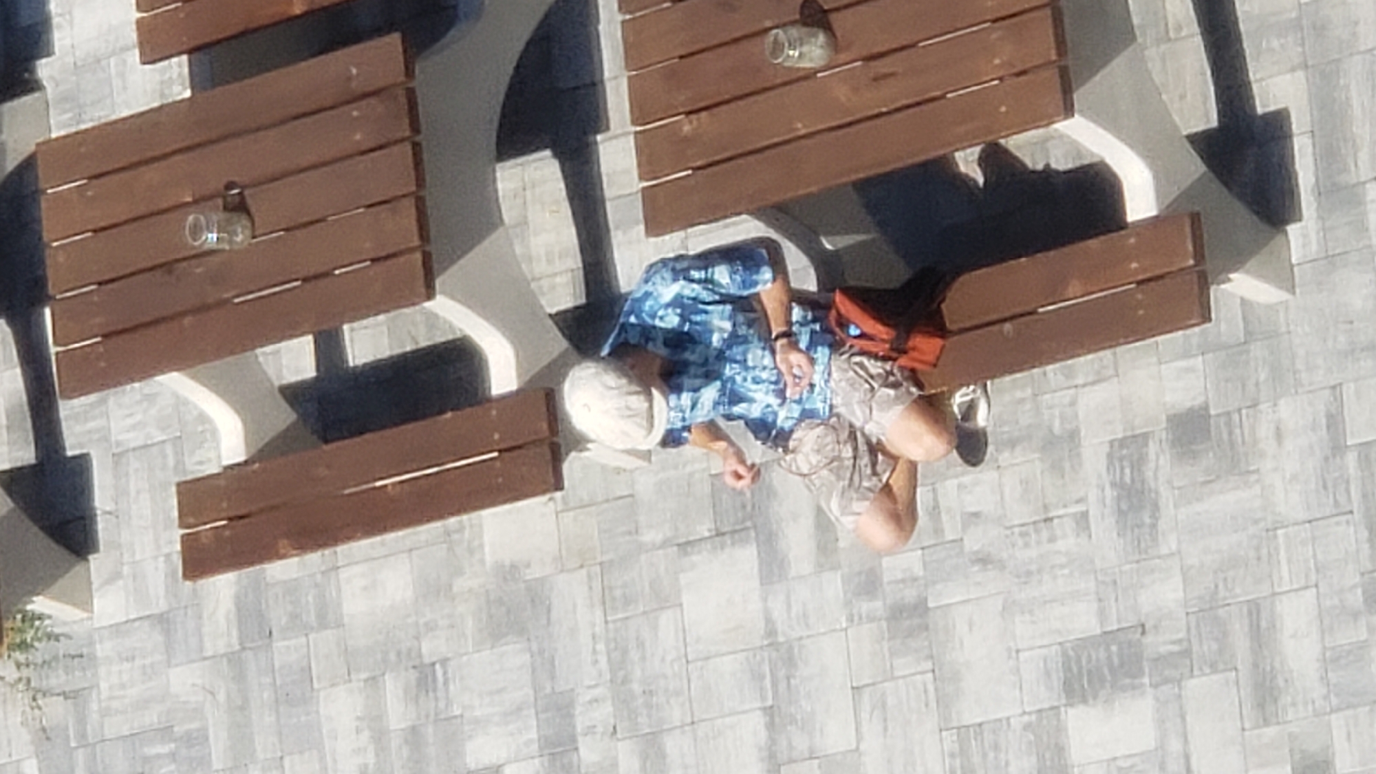 A person wearing a white hat, a blue and white patterned short-sleeved shirt, and patterned shorts is sitting on a bench at a picnic table. The person is holding something in their hands, and appears to be manipulating it. A small orange bag is on the bench next to them. The picnic table is made of wood and concrete. The ground is paved with gray and white square tiles. The scene is outdoors, likely in a park or public area. The image was taken from an overhead perspective. The location is identified as being in Prešov, Slovakia.