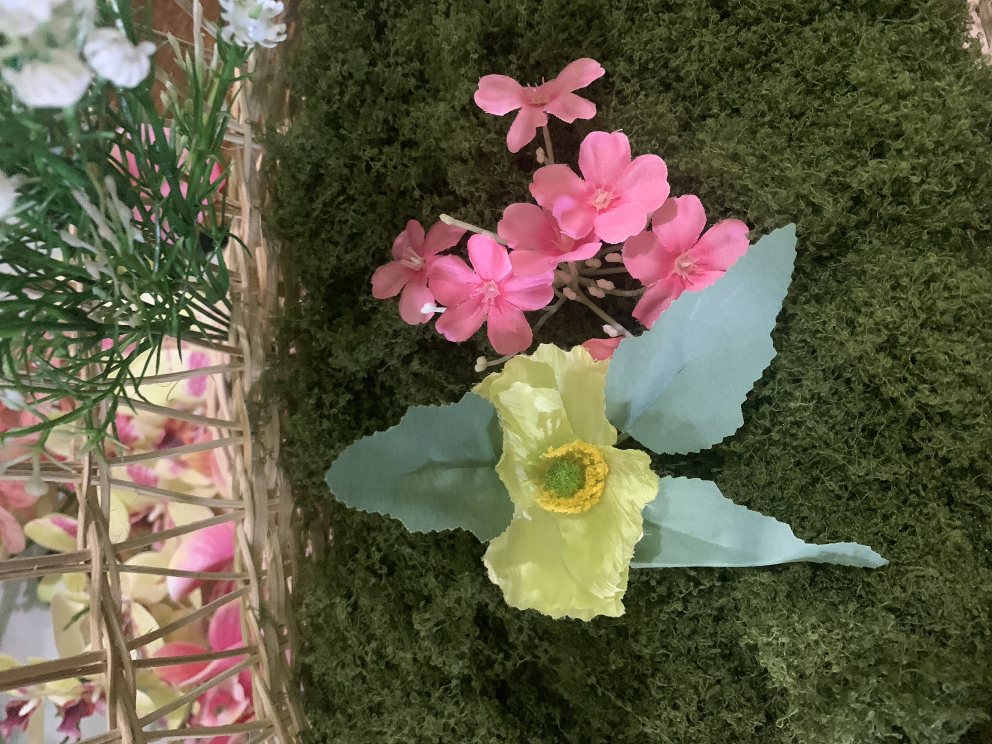 The image shows a close-up view of artificial flowers and greenery arranged on a textured green surface that resembles moss. In the upper portion of the image, a woven basket structure is visible, containing more artificial flowers in shades of pink and yellow, as well as green foliage. The focus is on a pale yellow flower with a vibrant yellow and green center, accompanied by two light blue-green leaves, placed on the moss. To the right of the yellow flower are several bright pink, small, five-petaled flowers clustered together. The scene appears to be a decorative arrangement, possibly for an event or display. There are no people or specific landmarks of Kuala Pembuang, Indonesia, visible in the image.