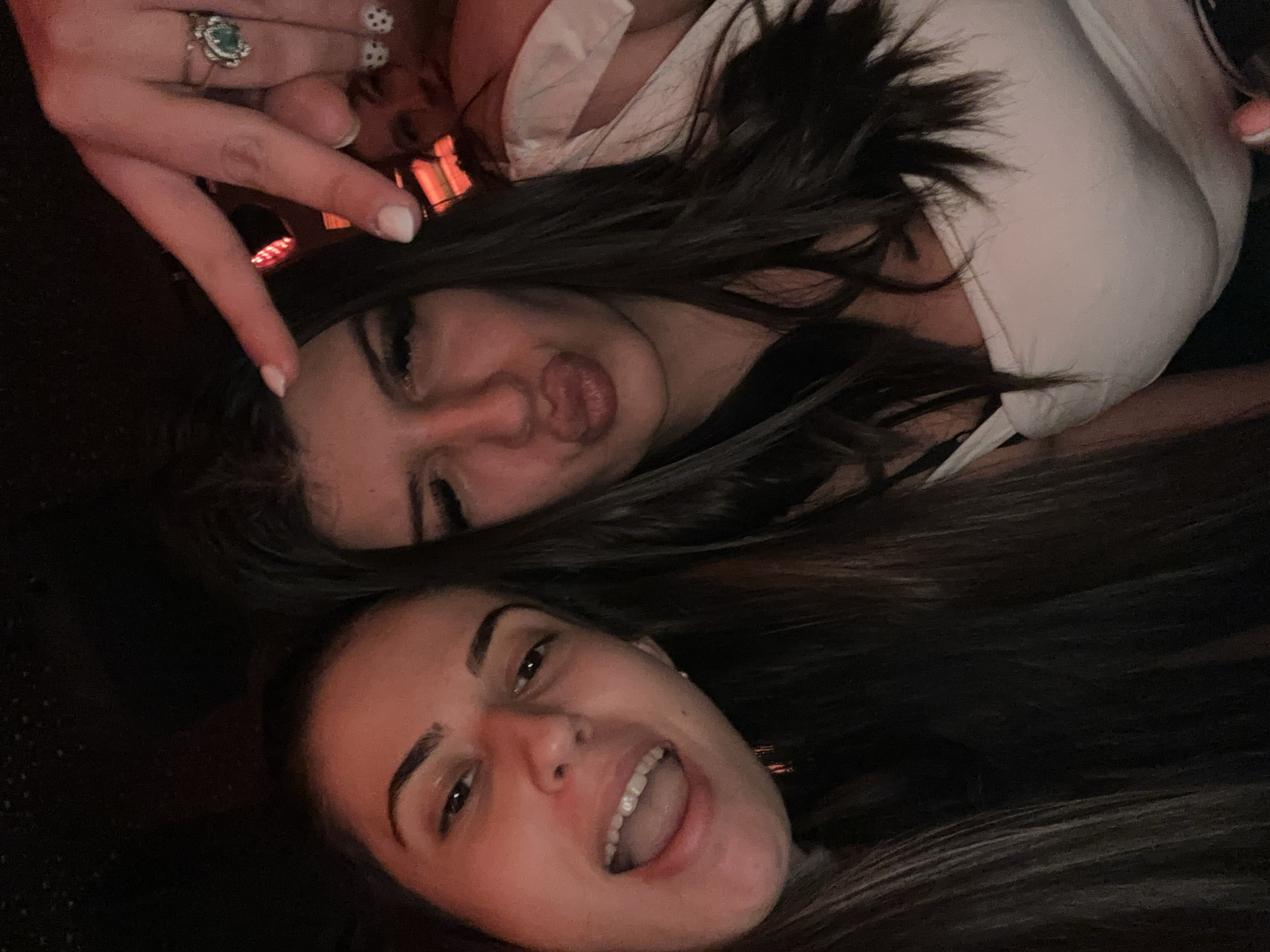 The image shows two young women lying down. One woman is puckering her lips and making a peace sign with her fingers which have white nail polish and an ornate ring on the ring finger. The other woman has her mouth open in what appears to be a smile or laugh. Both women have dark hair. The environment is dimly lit. Location: Unknown, Unknown.