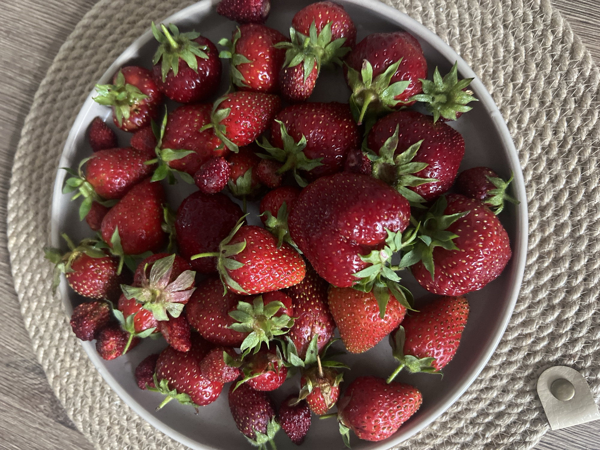 The image shows a plate of fresh strawberries on a woven placemat. The strawberries are a vibrant red color, and some have green stems and leaves attached. They vary in size, with some being quite large and others smaller. The plate is a light gray color, and the placemat is made of natural fibers, possibly jute or straw, in a neutral beige tone. The background appears to be a wooden surface. There are no people or animals visible in the image. The lighting suggests an indoor setting, possibly natural light coming from a window. The scene is focused on the strawberries, highlighting their texture and freshness. The image does not contain any specific information or visual cues that would allow for determining a location within Votkinsk, Russia.