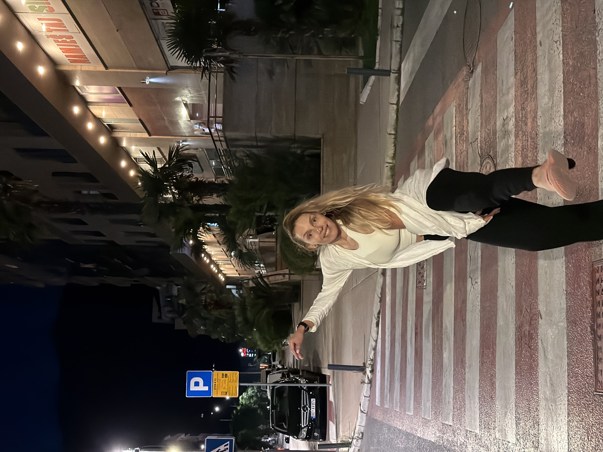 In Budva, Montenegro, a woman with long blonde hair, wearing a white top and jacket over black pants, stands on a pedestrian crossing at night. She is captured mid-motion, with her left leg extended and her right arm reaching out. The scene is illuminated by streetlights and lights from a building in the background, which features signage in Cyrillic script, including "НАМИЕШ ТАЈ SI". A black car with license plate "BD AV 874" is parked on the street to the left, next to a blue and yellow parking sign. Palm trees are visible, indicating a warm climate. The pedestrian crossing has red and white stripes.
