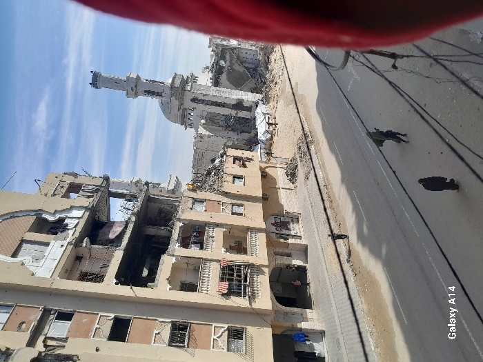 The image depicts a scene of extensive structural damage in Khan Yunis, Palestinian Territory, under a partially cloudy blue sky.
In the mid-ground, a multi-story, light-brown residential building occupies the left side of the frame, exhibiting severe damage. Multiple floors have exterior walls blown out, exposing interior rooms and structural elements, with debris hanging and partially collapsed balconies. Clothing is visible hanging from some of the remaining balcony railings. At street level, two adults and a child are visible near the base of this building on a paved sidewalk.
To the right and further back, the heavily damaged structure of a mosque is visible, featuring a white minaret that remains largely intact but with significant destruction around its base and dome. Rubble surrounds the mosque.
In the foreground and mid-ground, a paved road with white lane markings stretches across the frame. Three individuals are present on or near the road: one person in dark clothing walks on the left sidewalk; another person in dark clothing walks toward the viewer in the road's center, carrying a large, dark object on their back; and a third person in dark clothing walks away from the viewer on the right side of the road. Multiple overhead utility lines cross the upper part of the road.
Beyond the road and to the right, a cleared area contains piles of rubble and at least two white tent-like temporary shelters. The image is captured from an elevated perspective, looking down on the street and damaged structures. A portion of a red fabric object partially obstructs the view on the far right side of the frame. A "Galaxy A14" watermark is present in the lower-left corner.