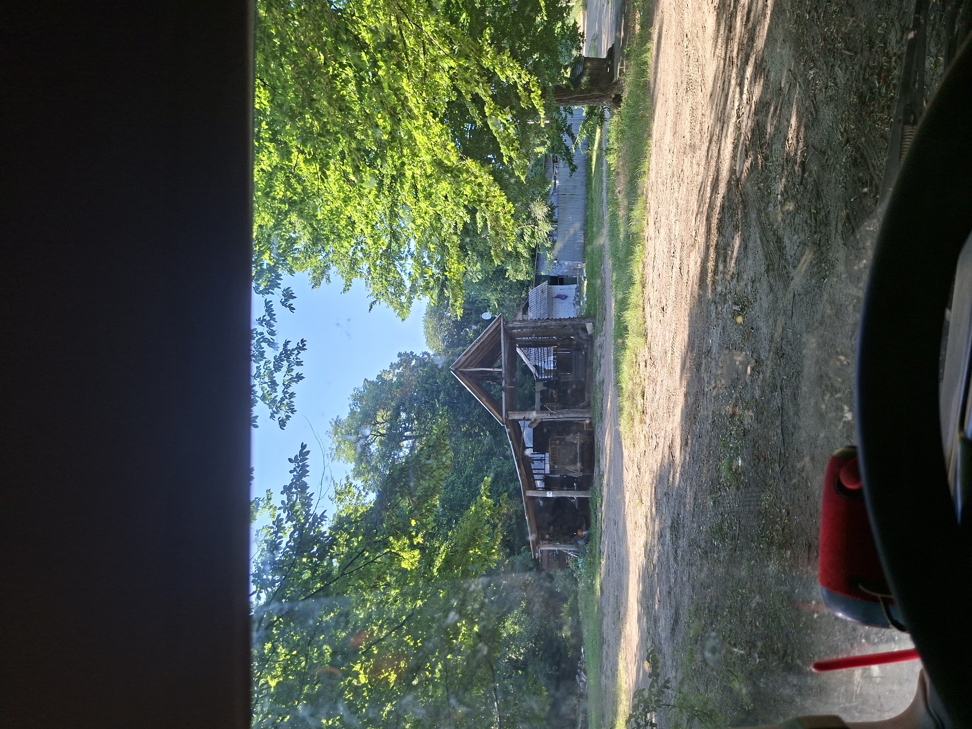 The image shows a view from inside a vehicle, looking out at a rustic wooden structure. The structure appears to be a shed or a small barn, with a pitched roof and open sides, revealing some items stored within. The building is situated in a rural setting, surrounded by lush green trees. A dirt road runs alongside the structure, with tire tracks visible on its surface, suggesting recent vehicle activity. Sunlight filters through the leaves of the trees, casting dappled shadows on the ground. The sky is a clear blue. On the right side of the frame, a part of the vehicle's interior is visible, including the steering wheel and a red object, possibly a seat or a piece of equipment. The scene is set during the daytime, with bright sunlight indicating fair weather. The overall impression is one of a peaceful, natural environment with a touch of rustic charm. The image was taken in Suruceni, Moldova.