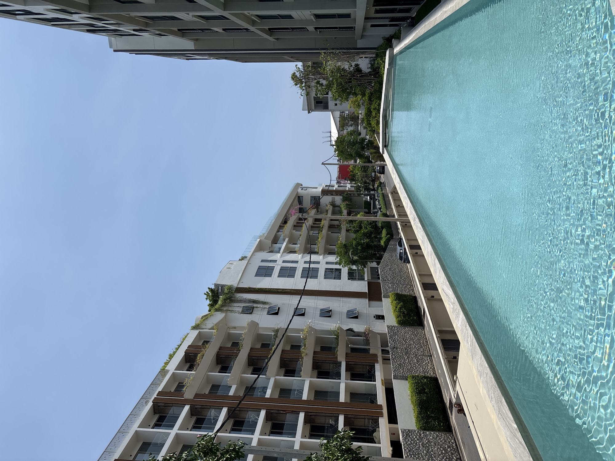 The image shows a swimming pool on a rooftop in Choeng Thale, Thailand. A tall, modern apartment building is visible to the left, with balconies and greenery. The clear blue sky is above. The building has white walls, with some windows open. A grey building can be seen in the distance.