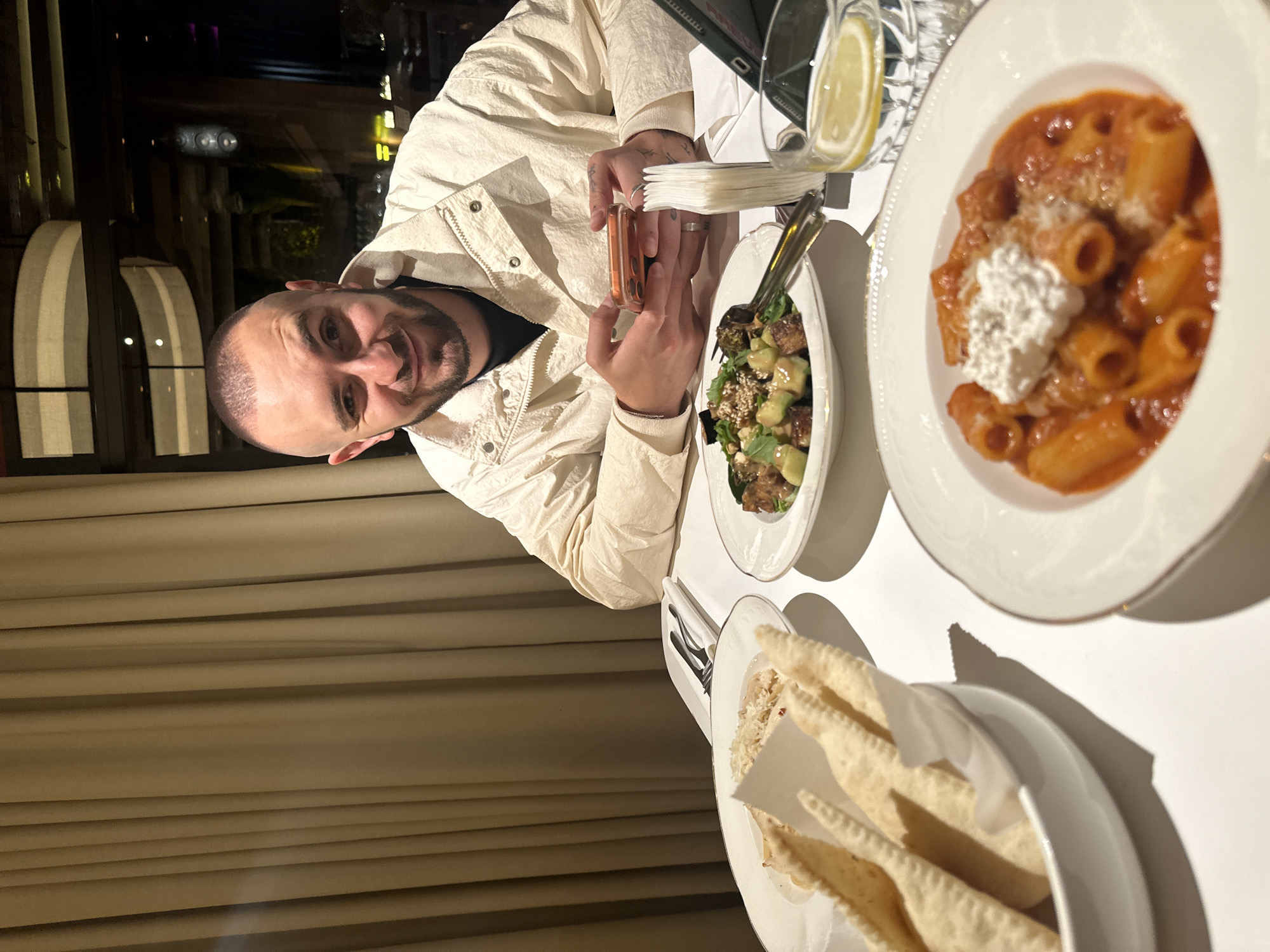 The scene depicts a male subject seated at a white-clothed dining table in an upscale restaurant in Yerevan, Armenia. The subject, an adult male with a closely-cropped haircut and a dark beard, is wearing a light-colored, possibly off-white, collared jacket over a dark shirt. He is holding a smartphone with an orange case in both hands, looking directly at the camera with a slight smile. A silver ring is visible on his left ring finger, and tattoos are present on his left wrist/hand.
The table is set with multiple dishes. In the foreground, a white plate contains Rigatoni pasta in a red tomato sauce, topped with a dollop of white ricotta cheese and grated Parmesan. To the left, a white bowl holds several flatbread triangles, possibly pita chips or lavash, partially wrapped in white paper. Further back, another white plate features a salad composed of mixed greens, avocado cubes, what appear to be roasted eggplant pieces, and sesame seeds. A clear, textured glass filled with water and a lemon slice is positioned on the right. Cutlery, including forks and knives, rests on white napkins. A stack of white paper napkins and a dark-cased electronic tablet are also present on the table.
The environment is characterized by warm, dim lighting. The background on the left features floor-to-ceiling light brown/beige curtains with vertical pleats. Behind the subject on the right, large windows reveal a dark outdoor scene with blurred urban lights and modern architectural elements, including horizontal illuminated bands, indicating an evening setting.