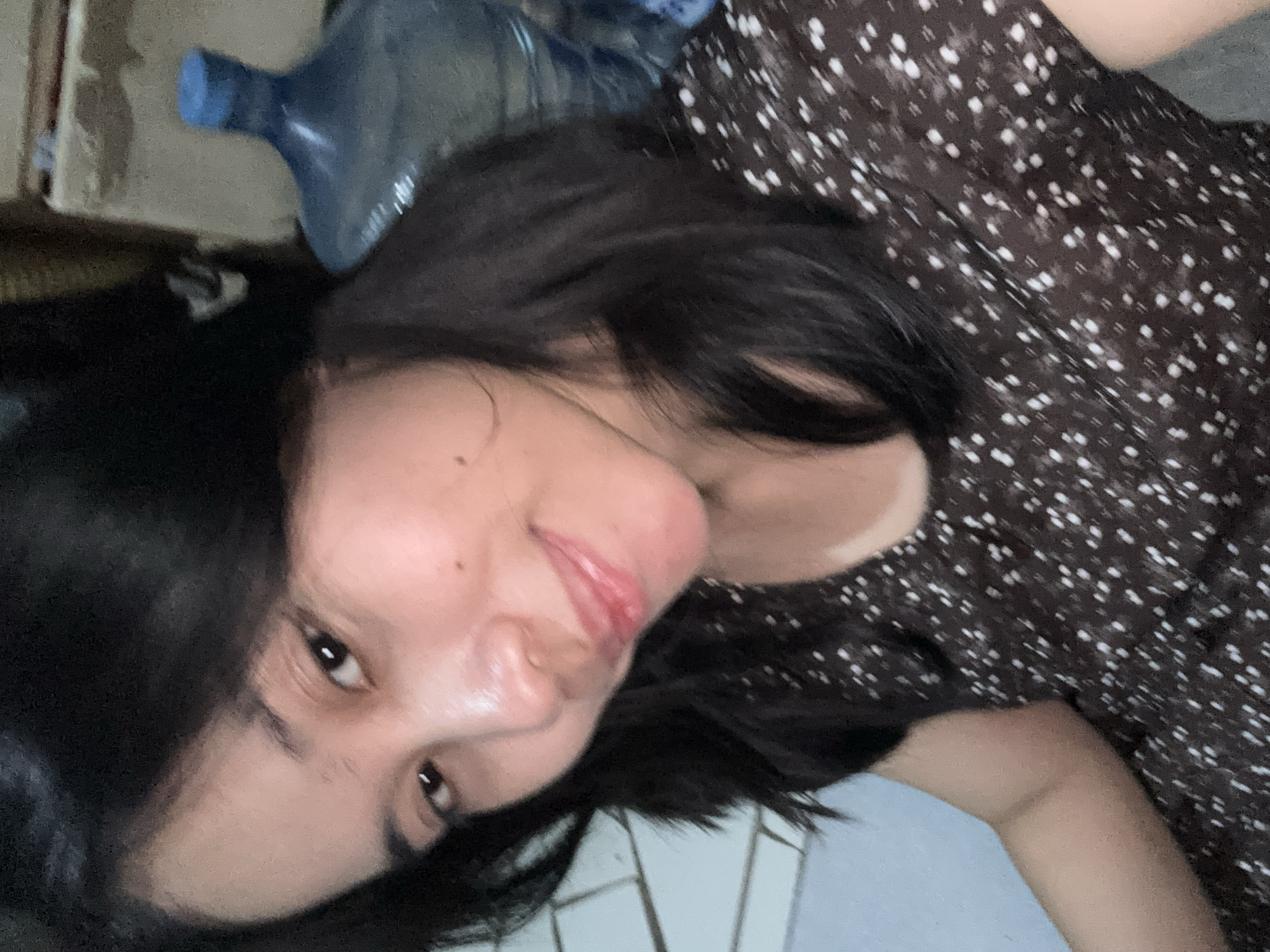 A young woman with dark hair is taking a selfie indoors. She is wearing a dark dress with a white polka dot pattern. In the background, a large blue water jug with a blue cap is visible. The woman is looking towards the camera and smiling. The lighting appears to be natural. The scene is likely a residential setting in East Jakarta, Indonesia.