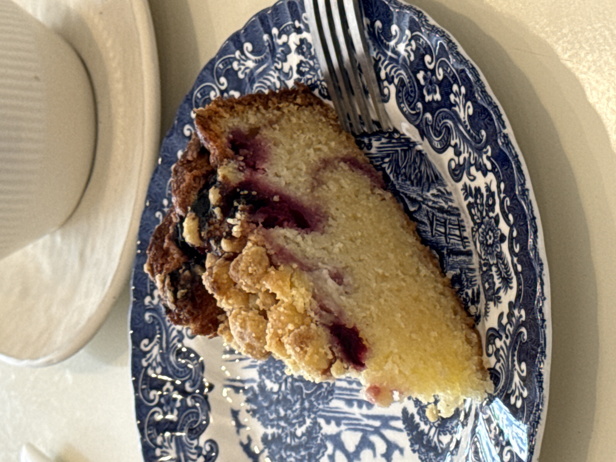 A slice of berry coffee cake with a crumb topping sits on a blue and white patterned plate. A fork rests on the right side of the plate. In the background, a white cup and saucer are visible. The scene is likely set on a table in Budva, Montenegro, perhaps at a cafe or restaurant.