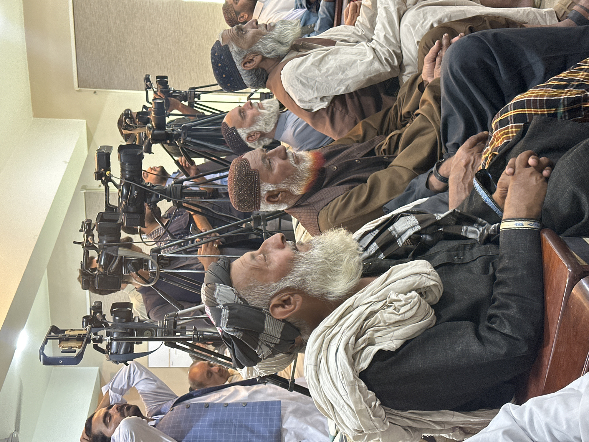 The image shows a group of men, likely elders, seated in a row, facing a bank of video cameras on tripods. The men are dressed in traditional attire, including turbans and shawls. The scene appears to be a formal setting, possibly a press conference or meeting, taking place indoors. The location is the city of Quetta City Tehsil, Pakistan.