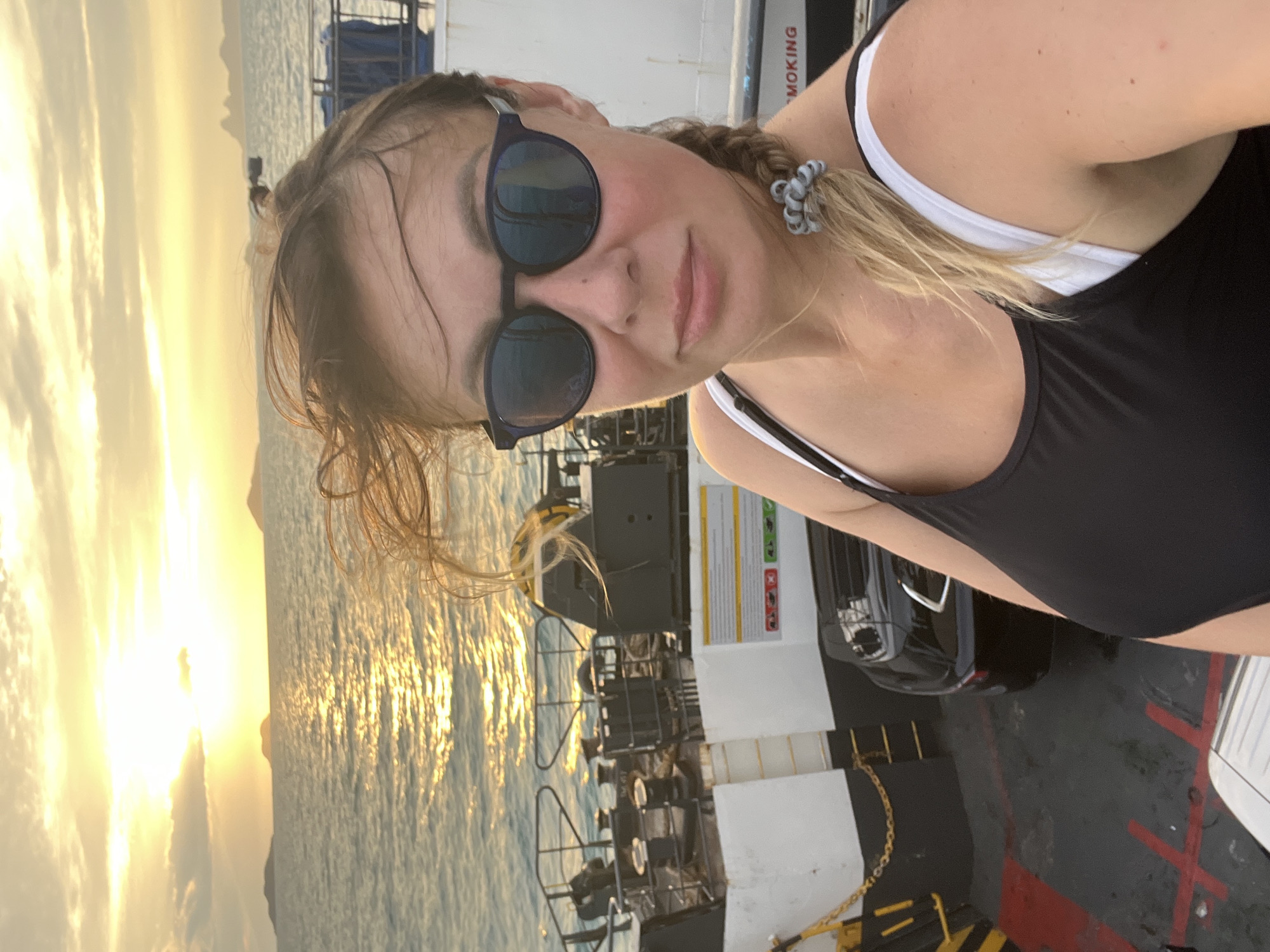 In a selfie taken on a ferry at sunset, a woman with blonde hair in a braid and wearing sunglasses and a black tank top over a white bra looks at the camera. The sun is setting over the ocean, casting a golden glow on the water and the sky, with islands in the distance. The ferry deck is visible behind her, with a black car parked on it and various pieces of equipment. A sign on the side of the ferry reads "SMOKING".