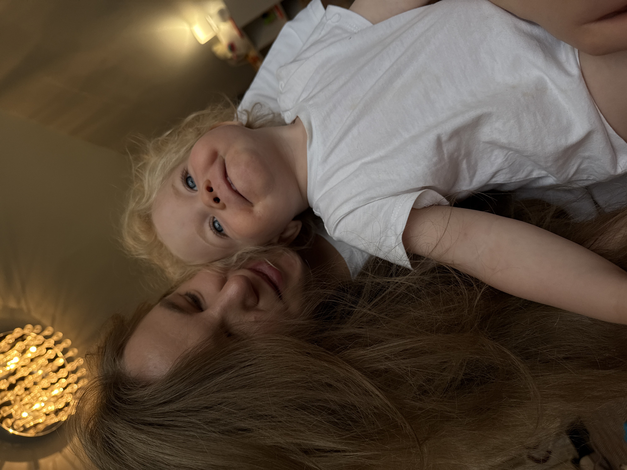 A close-up shot captures a moment between a woman and a young child, both wearing white t-shirts. The woman, with long, light brown hair, leans in towards the child, her eyes closed and a gentle smile on her face. The child, with curly blonde hair and striking blue eyes, looks upwards with a wide, joyful smile. The background is softly lit, with a chandelier visible in the upper left corner and a lamp on the right, suggesting an indoor setting. The scene appears intimate and tender, focusing on the connection between the two individuals. The specific location in Vladimir, Russia, cannot be determined from the visual information provided in the image.