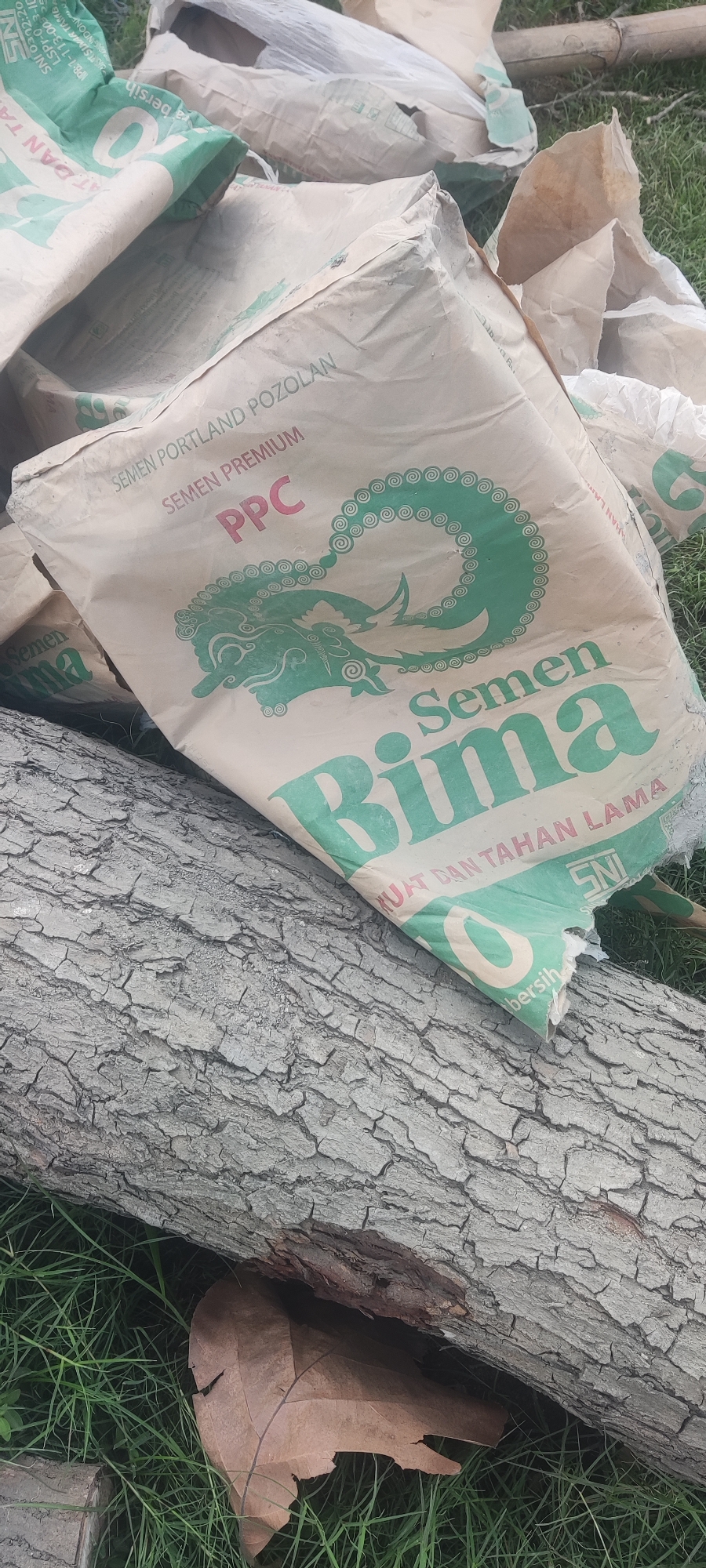The image displays several bags of cement, some of which are damaged, lying on the ground covered in grass. A large, rough-barked tree trunk is prominently featured in the foreground, angled diagonally across the frame. The cement bags are predominantly cream-colored with green lettering. One bag clearly shows the brand name "Bima" in large green letters, along with text that reads "SEMEN PORTLAND POZOLAN PREMIUM PPC". Another bag, partially obscured, also has "Semen Bima" visible. The environment appears to be outdoors, with green grass covering the ground and a hint of a wooden pole in the upper right background. The overall scene suggests a construction or storage area, possibly at a site in Gebangilir, Indonesia, given the context. There are no people visible in the image.