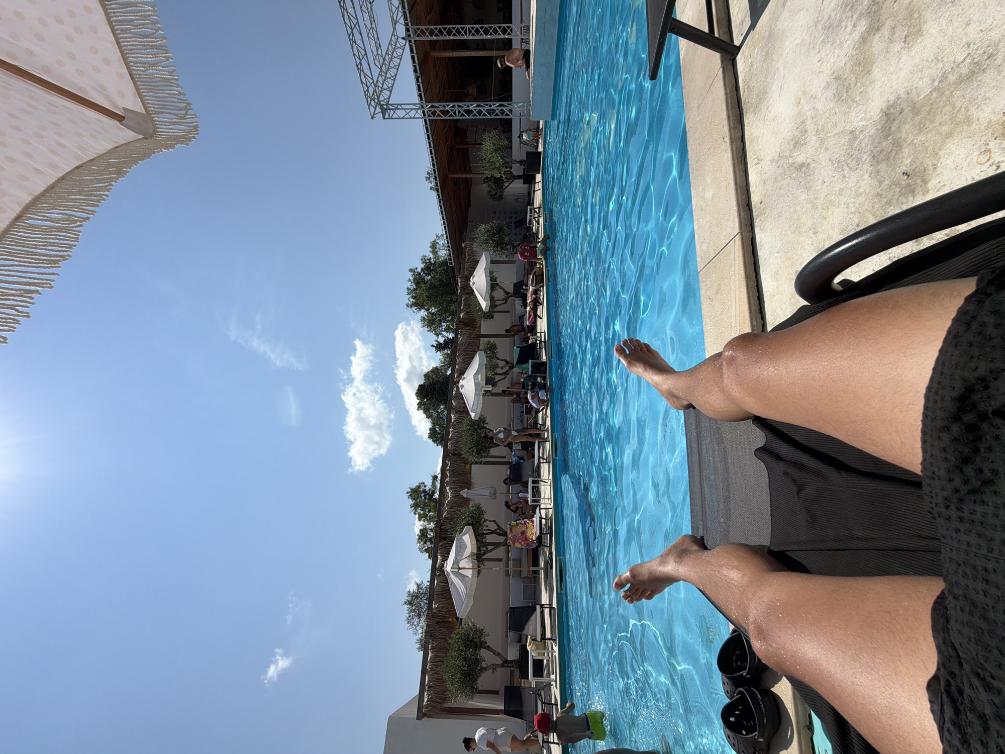 The image depicts a sunny day at a poolside resort. In the foreground, a person's legs are visible, stretched out on a dark recliner. Black Crocs are placed next to the recliner. The poolside is made of concrete, and a bright blue swimming pool is filled with clear water. Several people are seen relaxing by the pool on loungers or sitting on the edge. Some are in swimwear, while others are clothed. There are umbrellas providing shade, and trees with thatched roofs are present in the background. The sky is blue with a few white clouds, and the sun is bright, casting shadows. The location is identified as being in the city of Burgas, Bulgaria.