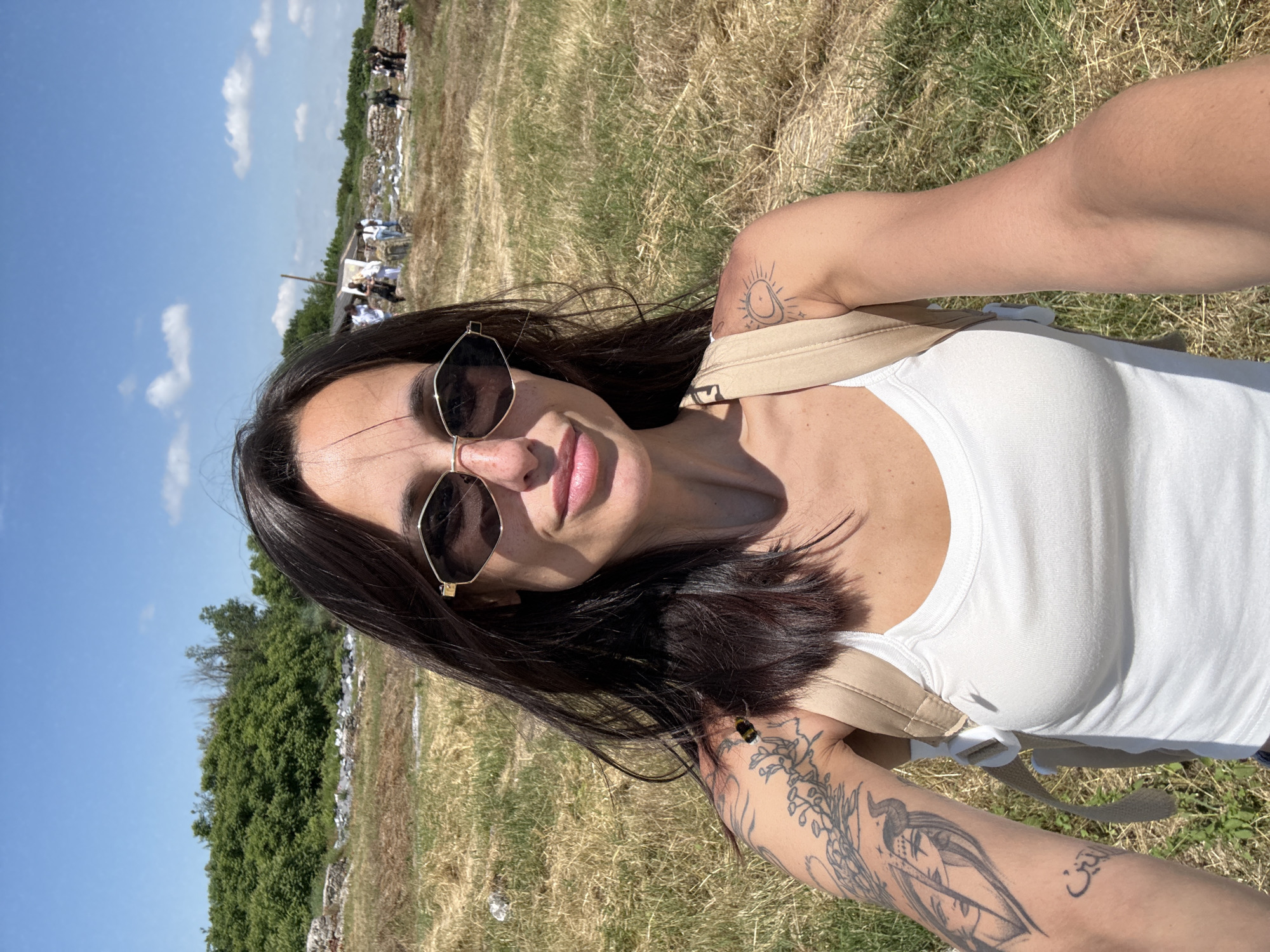 A female subject is captured in a front-facing selfie outdoors under bright sunlight. She is positioned centrally, wearing a white ribbed tank top and dark, hexagonal-shaped sunglasses with gold frames. Her long, dark hair is parted in the middle, with some strands appearing windblown. A subtle, closed-lip smile is evident. Freckles are visible on her nose and cheeks. Beige backpack straps are present over both shoulders.
Distinct tattoos are visible on her arms: on the upper left arm, an intricate design features a stylized female face with horns, integrated with botanical elements, and Arabic script below; on her right shoulder, a tattoo depicts a crescent moon alongside a sun.
The background reveals an expansive, dry, grassy field with areas of sparse vegetation. In the mid-distance to the left, a dense line of green trees and bushes is visible. Throughout the mid-ground and background, various stone ruins or structural remnants are scattered, particularly on the left and extending towards the center-right. Several individuals (approximately five to eight) are discernible in the far background, standing or moving near these structures. The sky is clear and blue with scattered white cumulus clouds. The scene is brightly lit, indicating a sunny day. The location is in Gigen, Bulgaria.