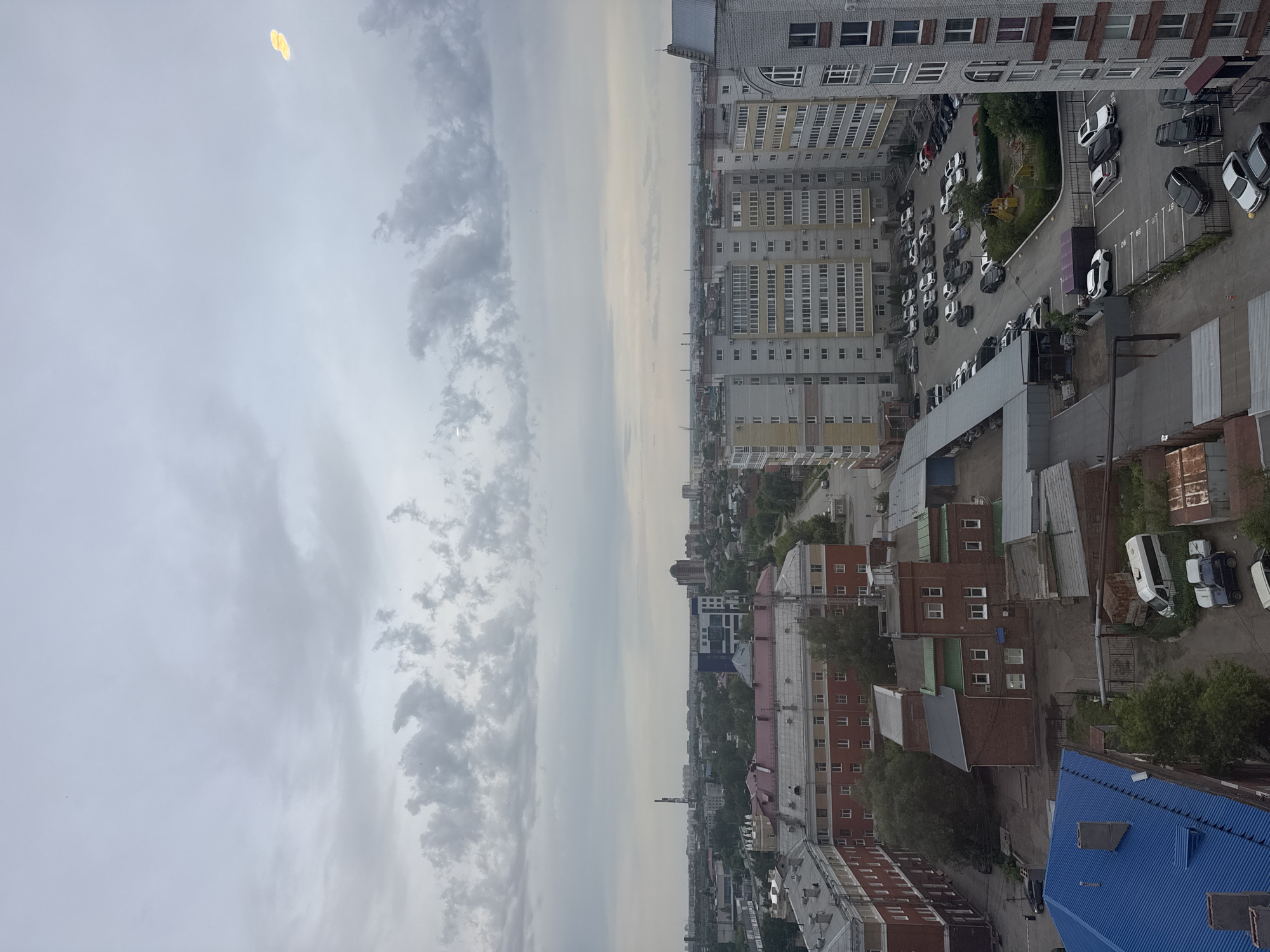 The image depicts an aerial view of an urban landscape under a cloudy sky, potentially in the city of Unknown, Unknown. The left side of the image is dominated by a sky with varying cloud formations and a yellowish light source. The right side reveals a cityscape, including tall apartment buildings with multiple windows and balconies, indicative of residential areas. A parking lot with numerous parked cars is visible next to the buildings. Lower buildings, potentially commercial or residential, with red brick facades and blue roofs can also be seen in the lower part of the image. The overall environment suggests a densely populated urban area.