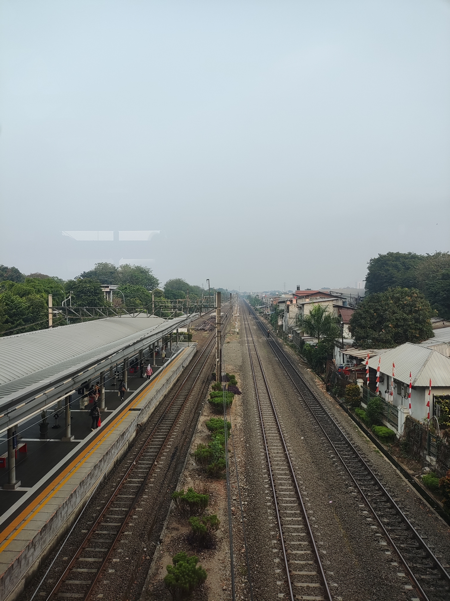 The image captures a view of a train station in East Jakarta, Indonesia, likely near a residential area. In the foreground, a covered train platform runs along two sets of train tracks. Several people are visible on the platform, some standing, others sitting, and one person appears to be walking. The platform is bordered by a yellow line and a concrete wall. The train tracks extend into the distance, with gravel ballast between the rails.
To the right of the tracks, a row of houses is visible, some with red and white flags displayed on their facades, suggesting a possible national celebration or commemoration. The houses appear to be of varying architectural styles and some show signs of wear. Dense green trees are interspersed with the buildings and line the horizon.
The sky is overcast and hazy, creating a diffused light that softens the overall scene. The atmosphere suggests a typical day in a densely populated urban environment. The image was likely taken from an elevated position, possibly from a bridge or another train.