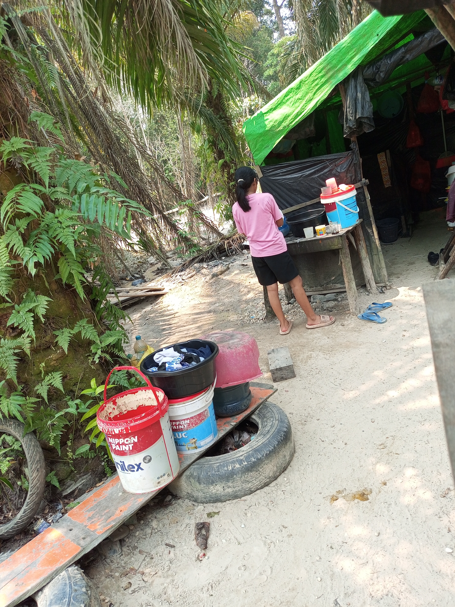 This image depicts an outdoor scene in Keling Panggau, Indonesia. The setting appears to be a makeshift shelter constructed with a green tarp for a roof and black plastic sheeting for walls. In the foreground, a young person wearing a pink t-shirt and black shorts stands near a wooden table. On the table are several containers, including a blue bucket with a red lid, a white bucket with red lettering, and what appears to be a small container of liquid. To the left of the table, a red and white paint bucket labeled "NIPPON PAINT" and another bucket labeled "Hilex" are visible. A black plastic basin filled with laundry sits on top of a red bucket and a tire. In the background, lush green foliage and palm trees suggest a tropical environment. The ground is sandy, with scattered debris and a few pairs of flip-flops. The overall impression is one of a simple, possibly temporary, living or working space.