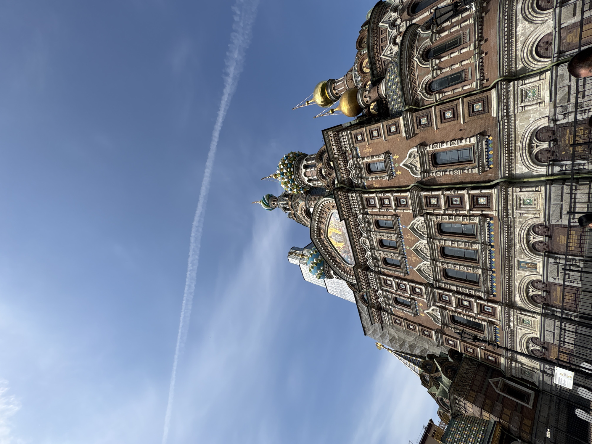The image depicts a low-angle view of the Church of the Savior on Spilled Blood in Saint Petersburg, Russia, taken during the day. The church's ornate facade, featuring multiple domes and detailed mosaic work, fills the right side of the frame. A clear blue sky occupies the left portion of the image, with a distinct contrail visible across it. A person's head is just visible on the upper-right edge of the frame. A metal fence can also be seen on the bottom right corner.