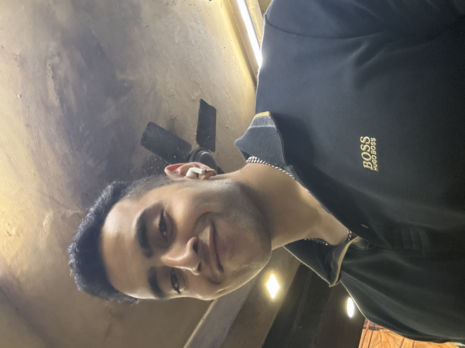 A young adult male is captured in a head-and-upper-torso portrait within an unspecified indoor location in Kolkata, India. He possesses dark hair, a light complexion, and is looking directly at the camera with a slight smile. He wears a dark blue or black polo shirt featuring a gold "BOSS HUGO BOSS" embroidery on the left chest and a silver-colored chain necklace. A white earbud is visible in his right ear, alongside a small gold-colored hoop earring. The background reveals a textured, light-colored ceiling with visible darker smudges or discoloration. A dark-bladed ceiling fan is partially visible mounted on the ceiling in the upper right quadrant, and a rectangular illuminated light fixture is present on the left side of the frame, indicating an artificially lit indoor environment.