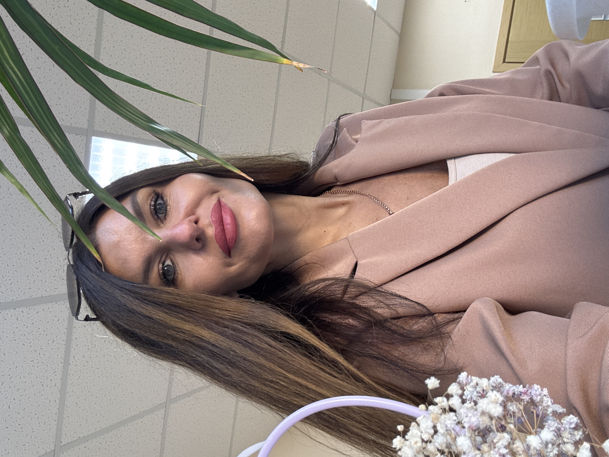 An over-the-shoulder, close-up, medium shot shows a woman with long, brown hair, wearing a light pink blazer over a white top and a silver necklace. She has on bold pink lipstick and sunglasses perched on her head. A plant with long, green leaves partially obscures the view on the right side of the frame, and a small bouquet of white flowers in a purple basket is visible on the left. The background consists of a white drop ceiling and a wall, suggesting an indoor setting. The image does not provide any information that would allow for location identification in Chaikovsky, Russia.