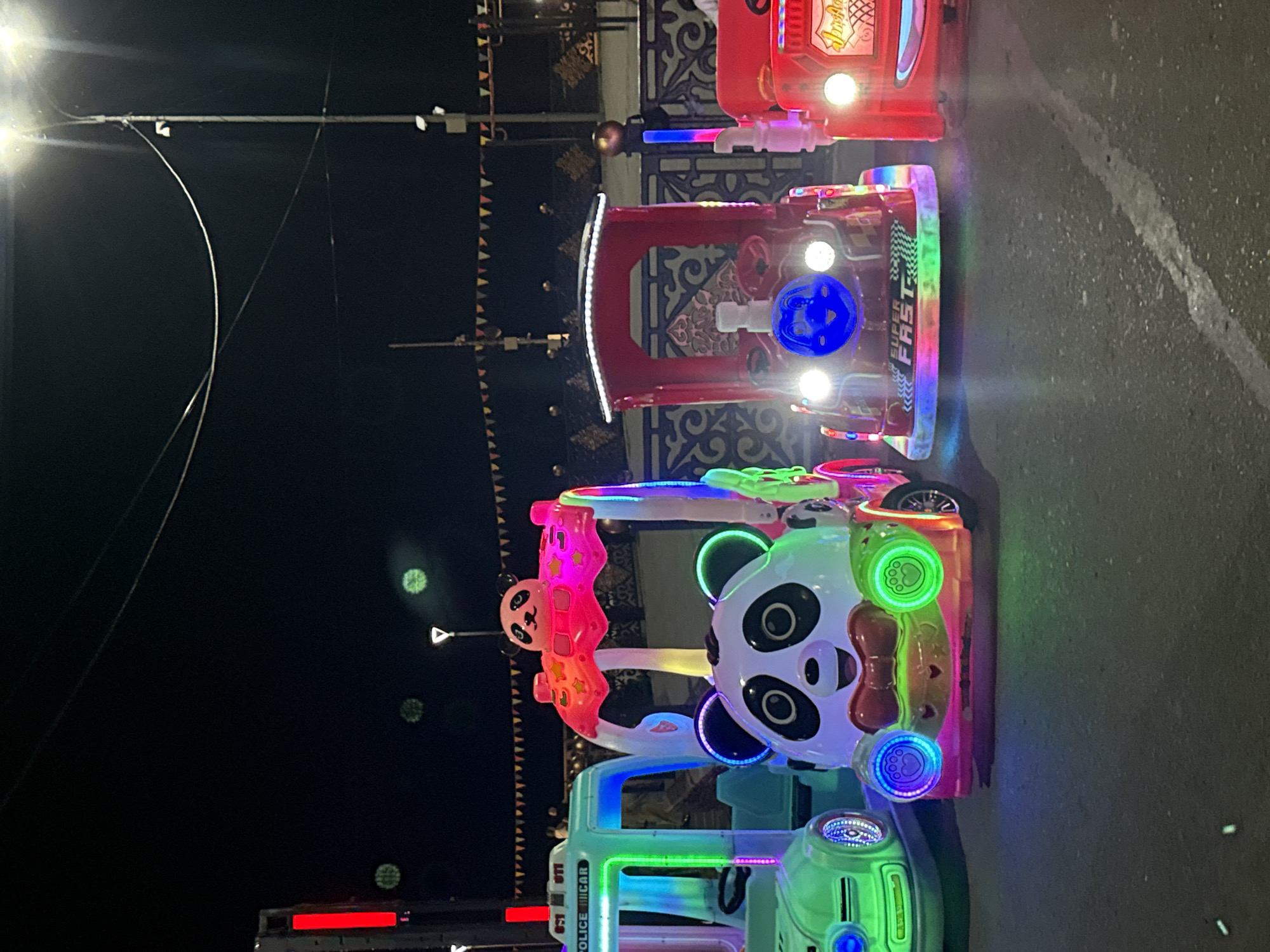 This is a nighttime, eye-level, medium shot of a row of illuminated kiddie rides in Pavlodar, Kazakhstan. From left to right, the rides are a green police car, a white panda-themed vehicle, a red train engine, and a red fire truck. The police car has a blue light bar and "POLICE" written on its side. The panda vehicle has a large panda face with colorful lights and a pink canopy. The red train has a blue face with lights for eyes and "SUPER FAST" written on its front. The red fire truck has "Ambulance" written on its side. The rides are parked on an asphalt surface with some light reflections. In the background, there are streetlights, wires, and a fence with decorative patterns. There are no people clearly visible interacting with the rides. The scene is illuminated by the rides themselves and streetlights.