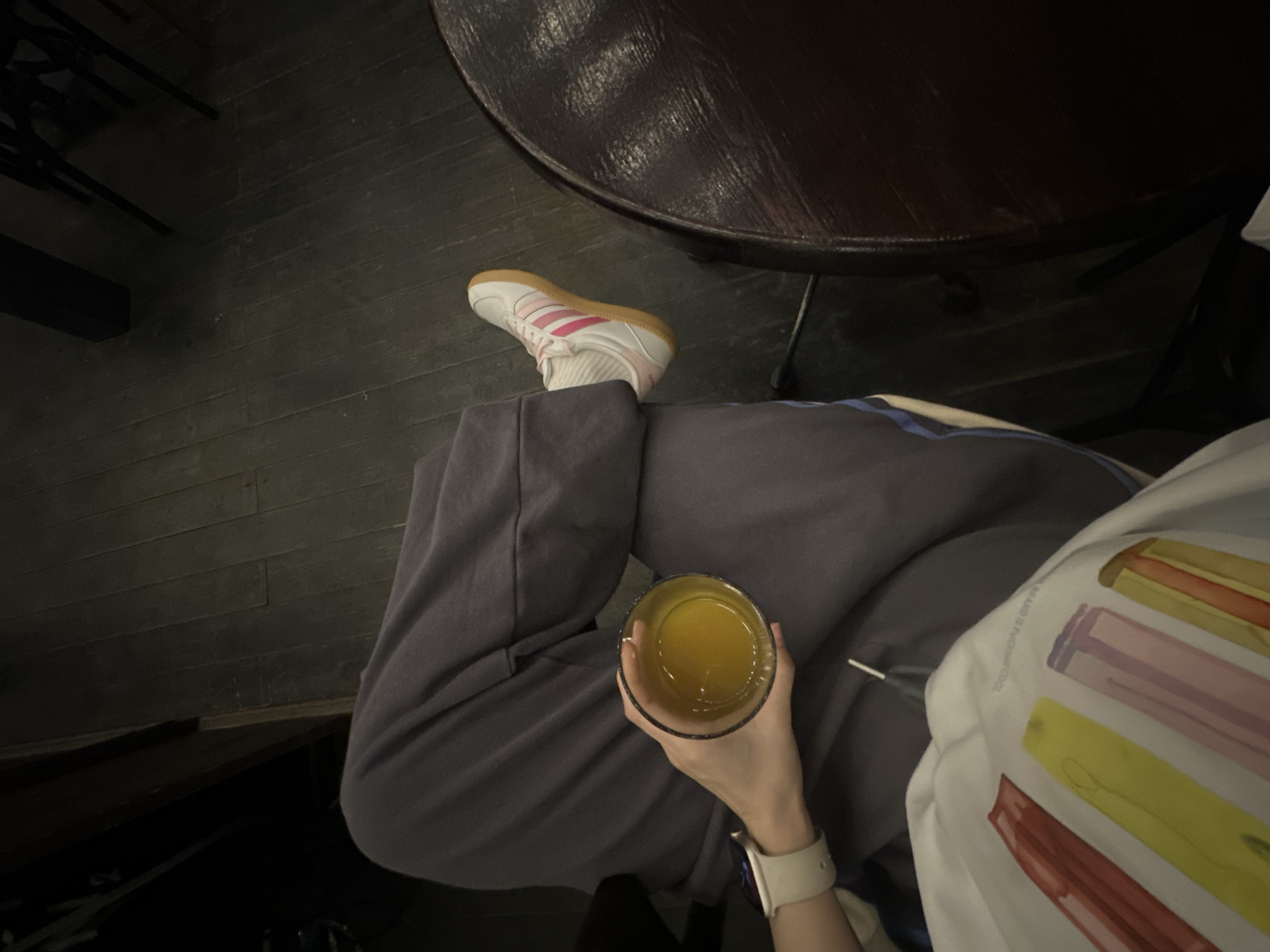 A person wearing gray sweatpants and a white shirt with colorful rectangular designs sits at a dark wooden table. The person is holding a glass of yellow liquid, possibly juice. The person is wearing white socks and Adidas Samba sneakers. The location is Perm, Russia.