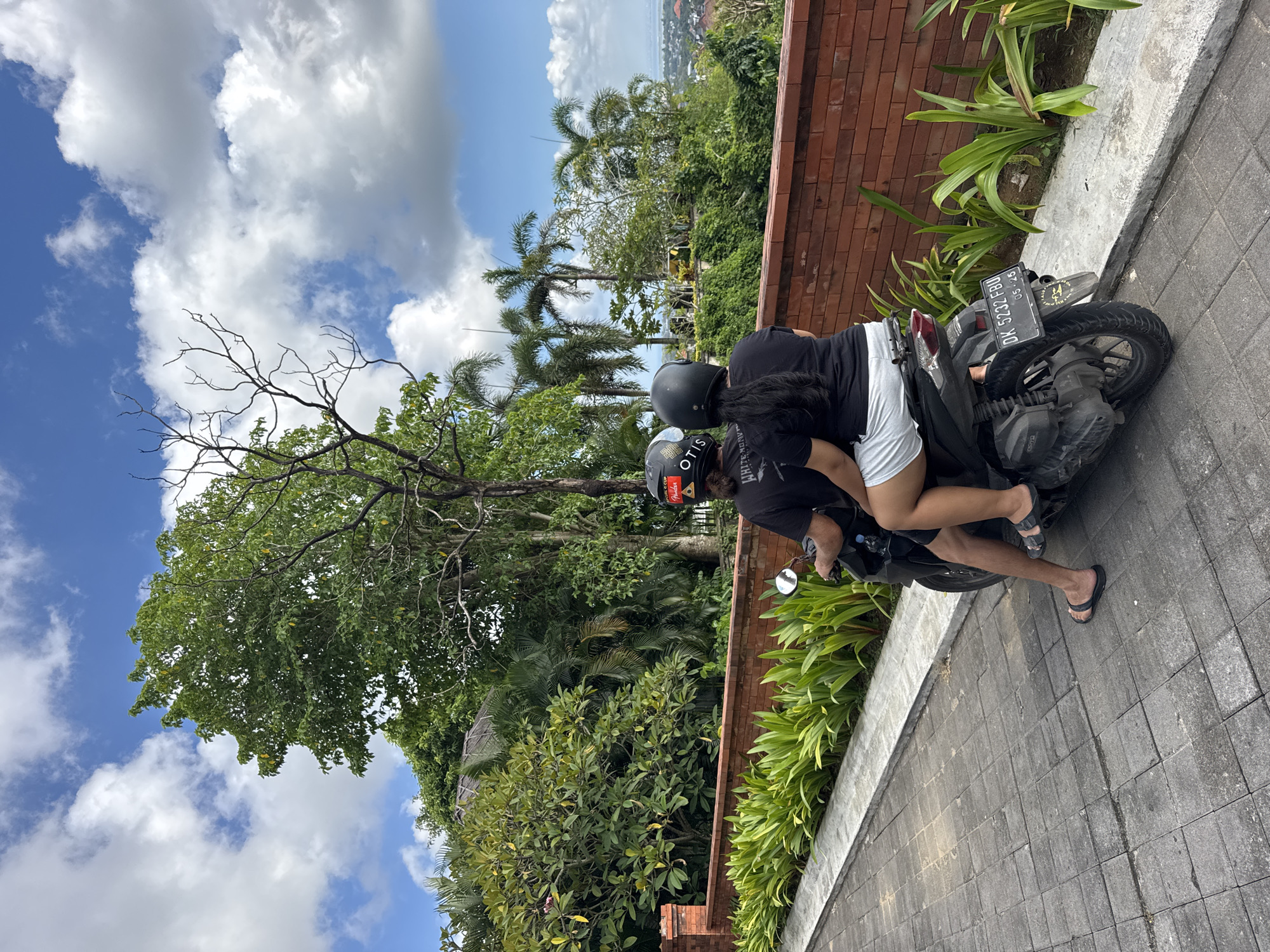 The image displays two individuals on a black scooter, situated on a paved path under a bright daytime sky in what appears to be a tropical setting in Kuta Selatan, Indonesia.
Both people are wearing helmets; the driver's helmet is black, and the passenger's dark helmet features the word "OTIS" in white text. The driver wears a black shirt, while the passenger is dressed in a black shirt, white shorts, and black flip-flops. The scooter's rear license plate is visible, showing "DK 5432 FBU" and "05 25", which likely indicates a Bali registration expiring in May 2025.
To the right of the paved path, a reddish-brown brick wall runs along the edge, adorned with vibrant green plants at its base. The background is dominated by lush, dense tropical vegetation, including tall palm trees and various other green trees. In the far distance, a glimpse of a coastal town or developed area suggests an elevated viewpoint. The sky is bright blue with scattered large, fluffy white clouds, indicating clear and sunny weather. The scene conveys a sense of a relaxed journey in a scenic, warm environment.