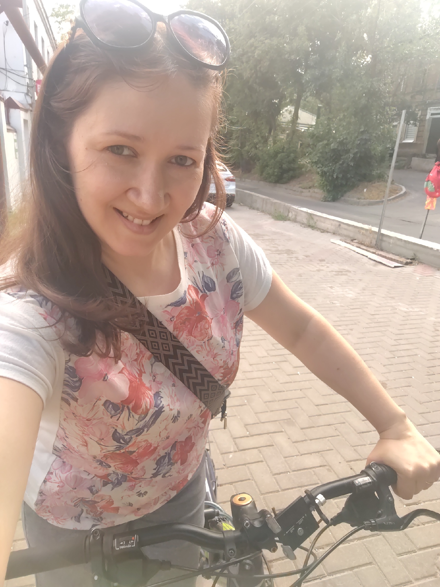 This is a selfie taken by a woman outdoors, likely on a bicycle. She is smiling and looking at the camera. She has brown hair and is wearing sunglasses on her head. Her t-shirt has a floral pattern with pink, red, blue, and white flowers. A patterned strap is visible across her chest. The background shows a paved walkway, some buildings, trees, and a street. The lighting suggests it is daytime, possibly sunny. The overall mood appears cheerful and relaxed. Visible text on the bicycle handlebars reads "1 2 3 4" and "Shimano".
