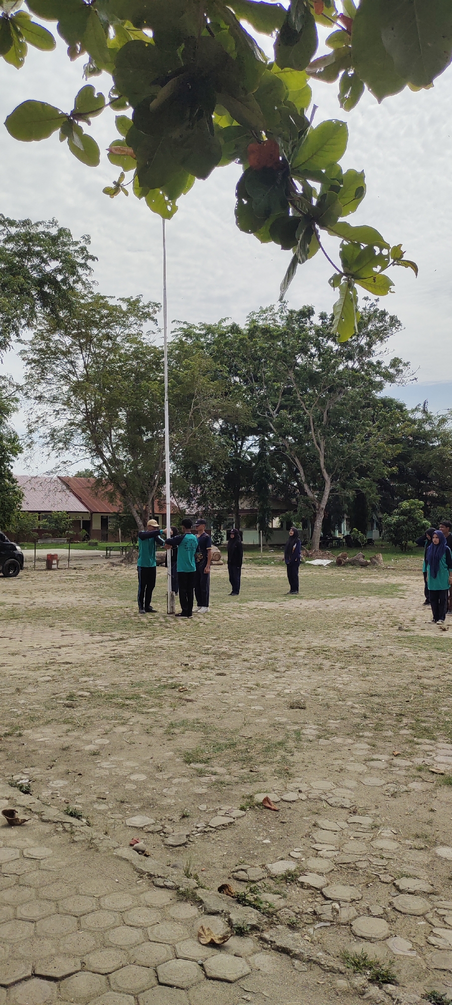 In Langnibong, Indonesia, a group of seven people, likely students and an instructor, are gathered around a flagpole in an outdoor, dusty field with scattered hexagonal paving stones. The individuals are dressed in uniforms or casual attire. Several are wearing teal shirts and dark pants, suggesting a school or organized group. One person in a dark uniform appears to be instructing or addressing the group. The background features several buildings with reddish roofs and lush green trees. The sky is overcast with white clouds. The scene appears to be set in a schoolyard or open area within the town.