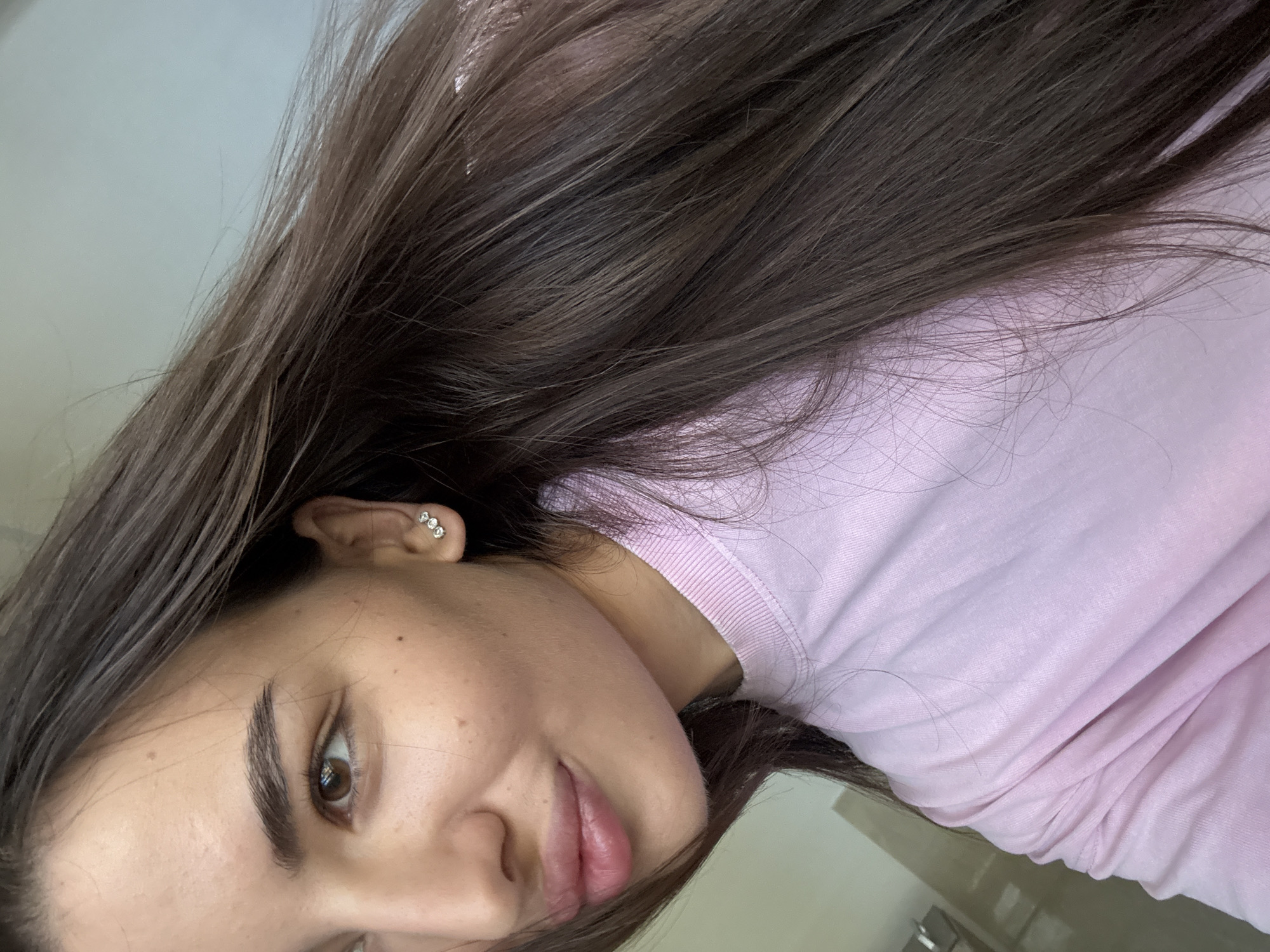 The image shows a close-up, low-angle outdoor shot of a young woman with long, dark brown hair and brown eyes. She is wearing a pink sweatshirt and a three-stone stud earring in her left ear. The background is blurred, but appears to be a light-colored wall, suggesting an urban setting. There is no specific indication of the location within Chișinău, Moldova.