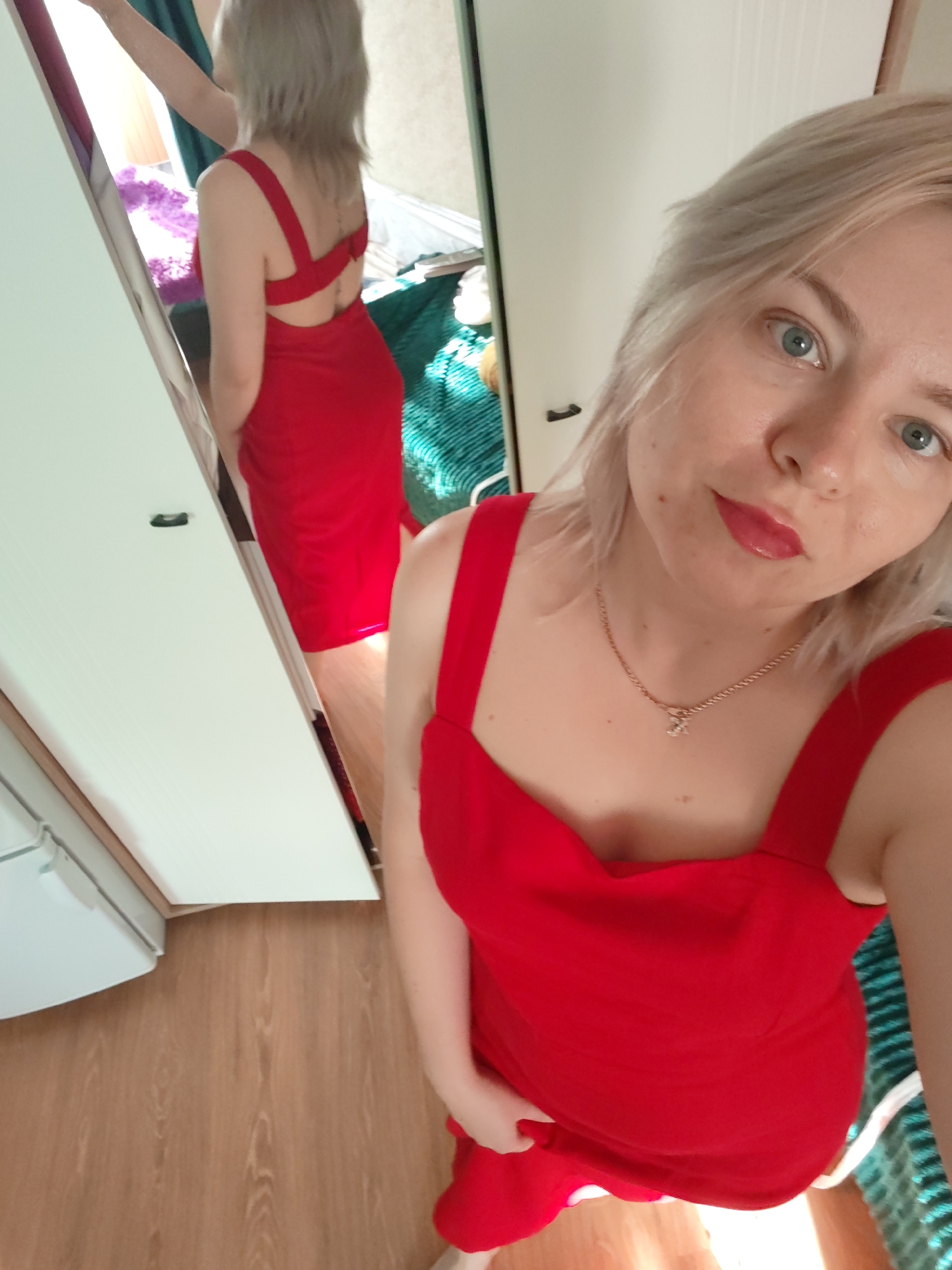 A blonde woman in a red dress is taking a selfie in front of a mirror. The reflection in the mirror shows the woman standing and facing away, revealing the back of her dress. The environment appears to be a room in a home, with a white door and wooden flooring visible. No information is available that indicates the city is Izhevsk, Russia.