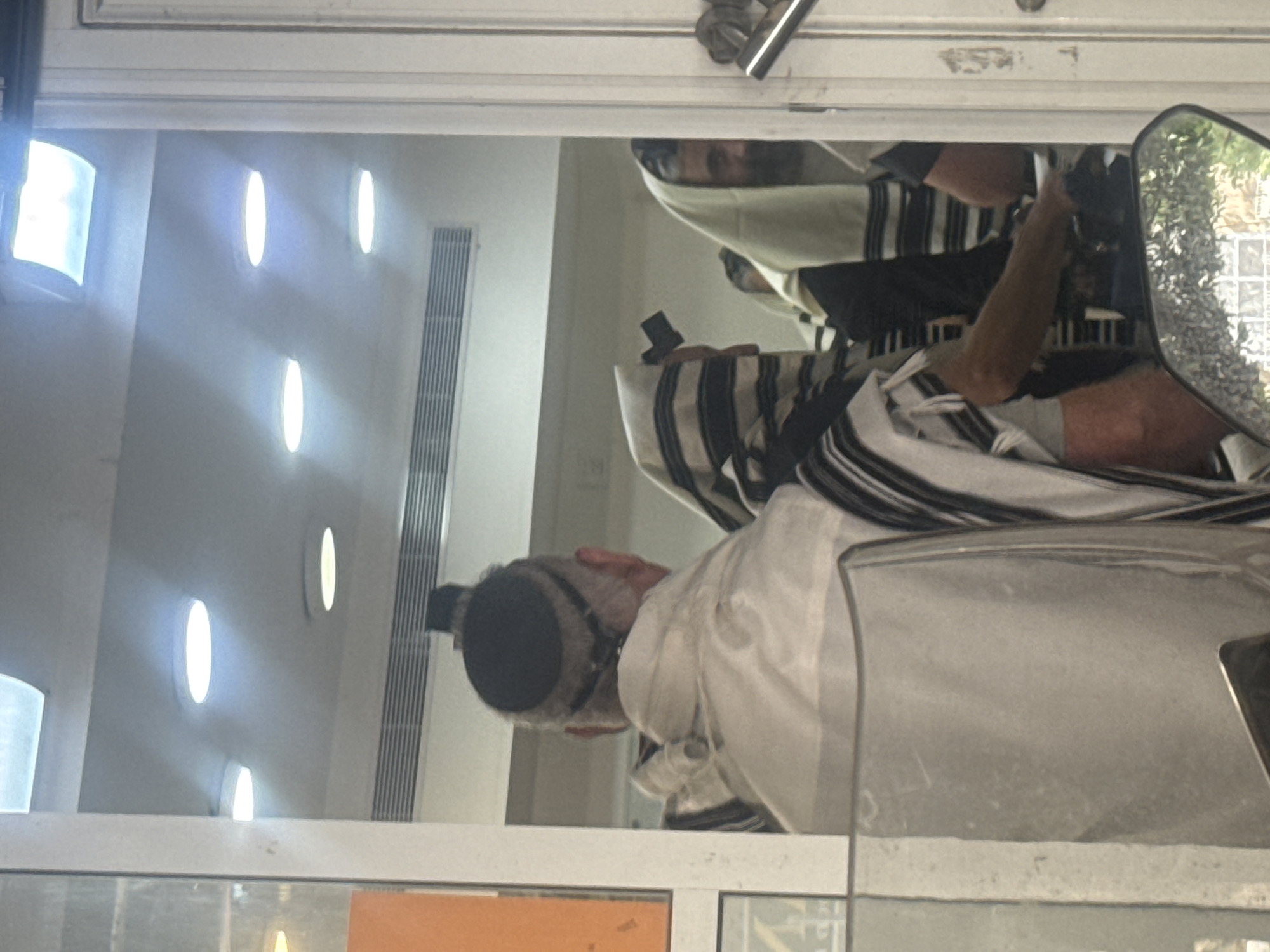 Reflection showing a man wearing a kippah and prayer shawl (tallit), with other men behind him also wearing prayer shawls. The location appears to be a window in a building with oval light fixtures and vertical air vents, reflecting the activity inside what is probably a synagogue in Givatayim, Israel.