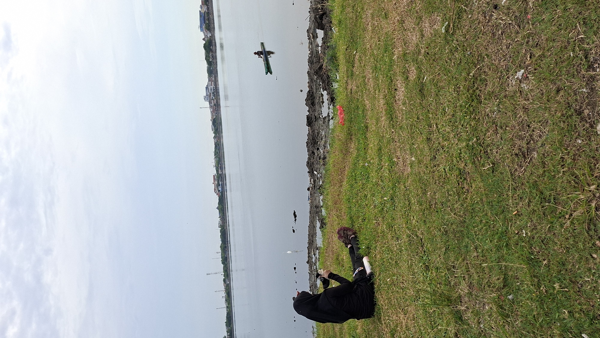A woman wearing a black hijab and black clothing sits on the grassy bank of a body of water, holding a camera. Her legs are crossed, and she is looking towards the water. In the distance, a person in a green canoe paddles across the water. The shoreline is littered with mud and debris. A white bird stands in the shallow water near the shore. The background shows a hazy cityscape with buildings and what appear to be telecommunication towers. The sky is overcast. The scene appears to be the water reservoir in the city of Lhokseumawe, Indonesia, possibly the reservoir on the west side of the city near the Unimal campus or the one near the Lhokseumawe Islamic Center.