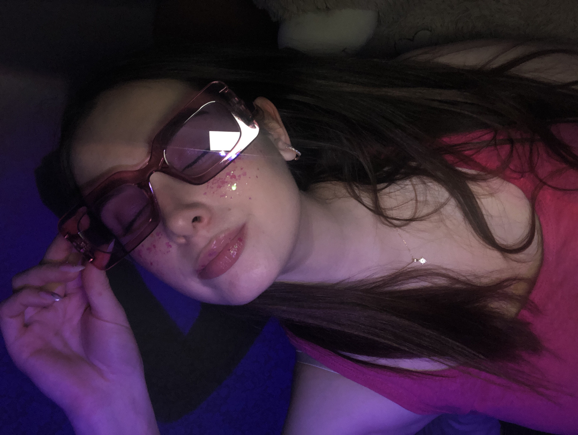 A young individual with long, dark brown hair is captured in a close-up, tilted portrait, lying down indoors. She wears large, pink-tinted rectangular sunglasses that obscure her eyes. Playful pink and purple glitter is scattered across her right cheek, mimicking freckles. Her lips are adorned with a shiny, light pink gloss. She is dressed in a fuchsia or magenta-colored top, and a delicate gold chain necklace with a small, sparkling pendant is visible around her neck. Her left hand, featuring a silver-toned ring on her ring finger, gently holds the arm of her sunglasses. The setting appears to be a dimly lit room, with a soft purple ambient light illuminating the lower left side of the frame. The background is dark and indistinct, with a blurry, light-colored, fluffy object partially visible in the upper right corner, suggesting a cozy, personal space like a bedroom or lounge. The time of day is likely evening or night due to the low light. The image conveys a cool, playful, and relaxed mood. No visible text is present.