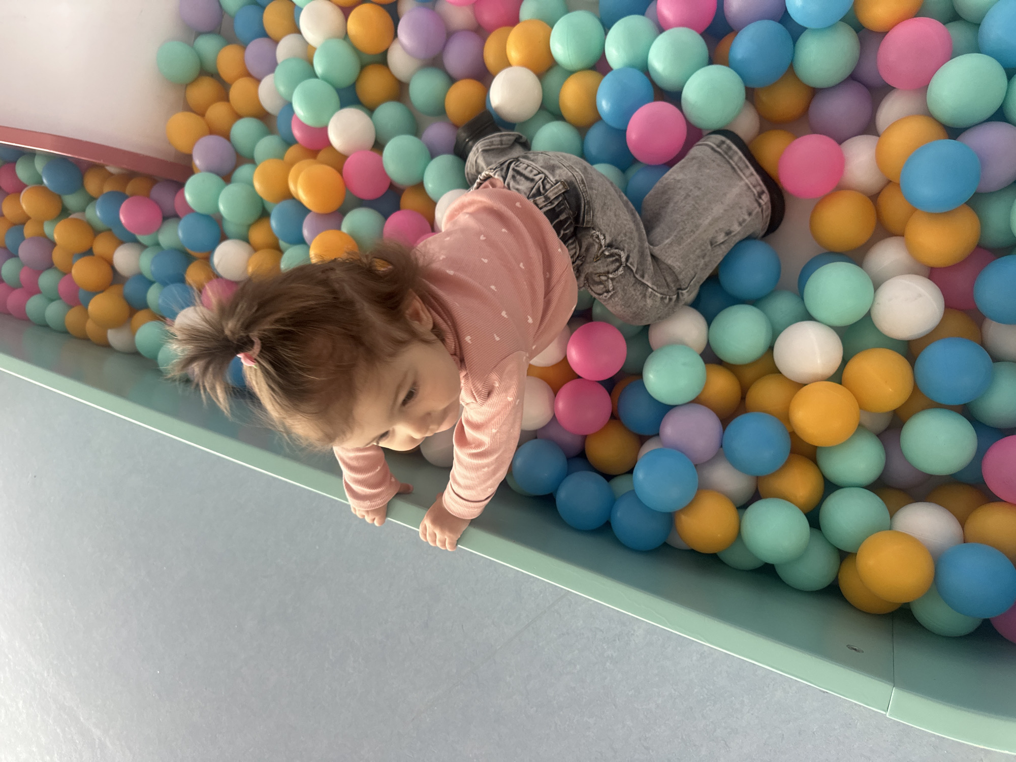 An image, rotated 90 degrees clockwise, captures a young child actively engaged in a colorful ball pit in Khimki, Russia. The child, a toddler with light brown hair tied back with a small pink hair tie, is dressed in a light pink long-sleeved top with a subtle dotted texture and grey denim jeans. One dark shoe is visible on their right foot.
The child is positioned with their upper body over the light green rim of the ball pit, hands extended onto the edge as if crawling or attempting to climb out or in. Their gaze is directed downwards, suggesting focus and curiosity in their activity.
The ball pit is densely filled with numerous plastic balls of uniform size in a soft, pastel color palette, including shades of mint green, light blue, yellow, pink, purple, and white. These balls largely obscure the child's lower body. Below the visible green rim of the ball pit, a light blue or grey floor or wall surface is seen. The background above the ball pit shows a white ceiling and an adjacent wall, indicating an indoor play environment. There is no visible text in the image.
