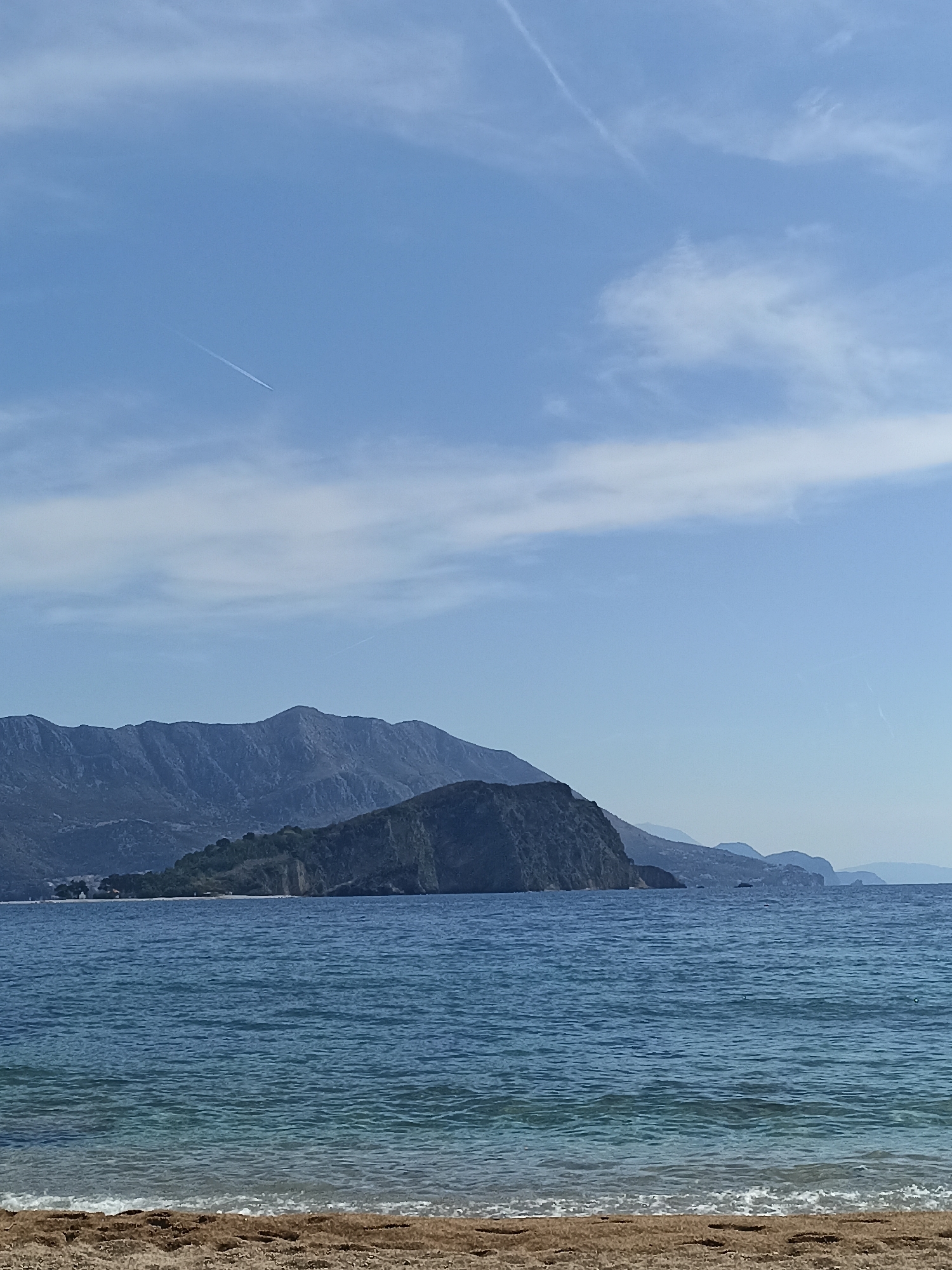 The image shows a coastal scene. A body of water, likely the sea, occupies the majority of the frame. In the background, there are mountains and a land formation resembling a large hill or small mountain extending into the water. The foreground consists of a sandy beach. The sky is blue with some cloud cover, and a contrail from an airplane is visible. The location appears to be an Unknown city within Unknown.